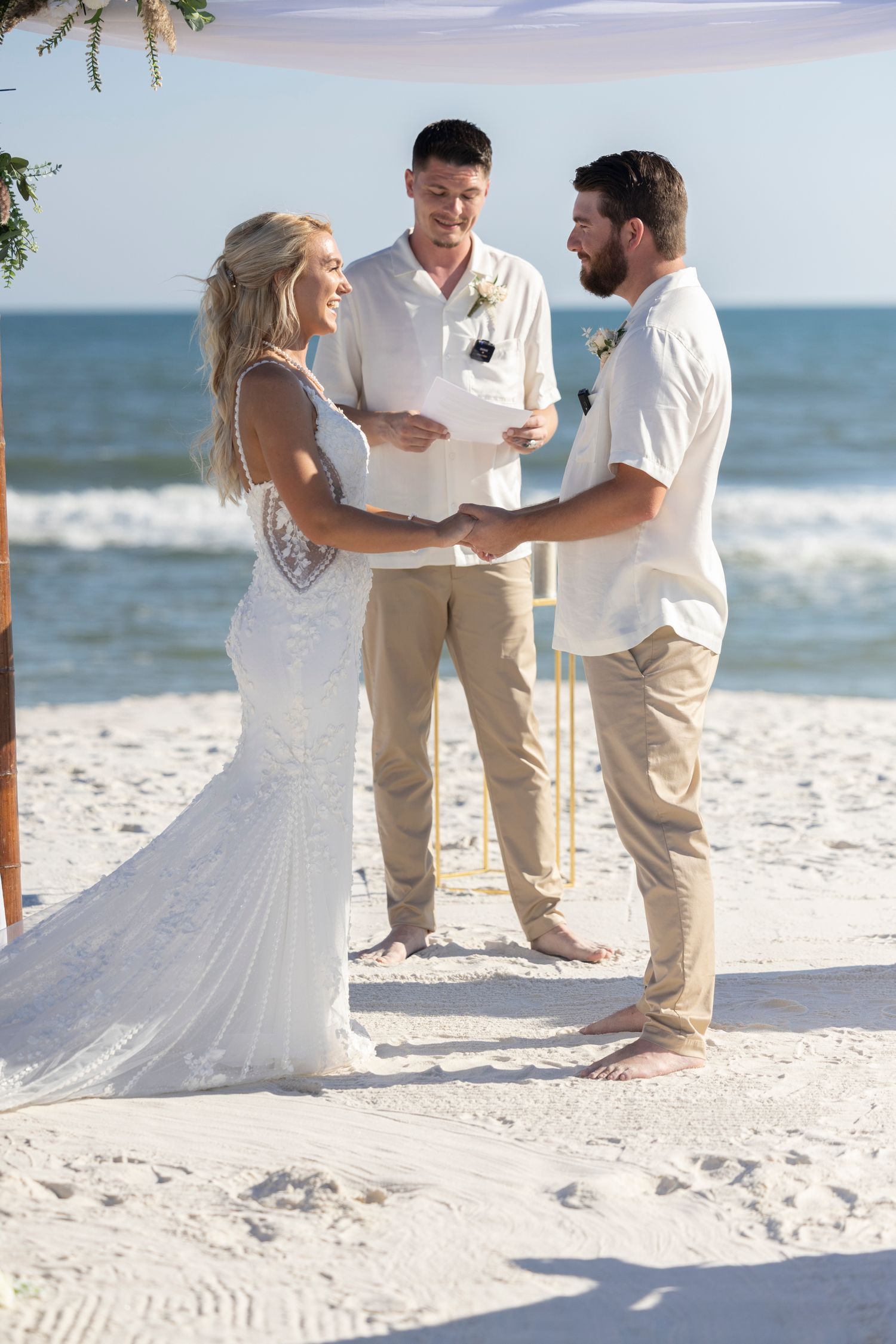 A beachside wedding ceremony takes place on white sand with ocean waves and palm trees in the background.