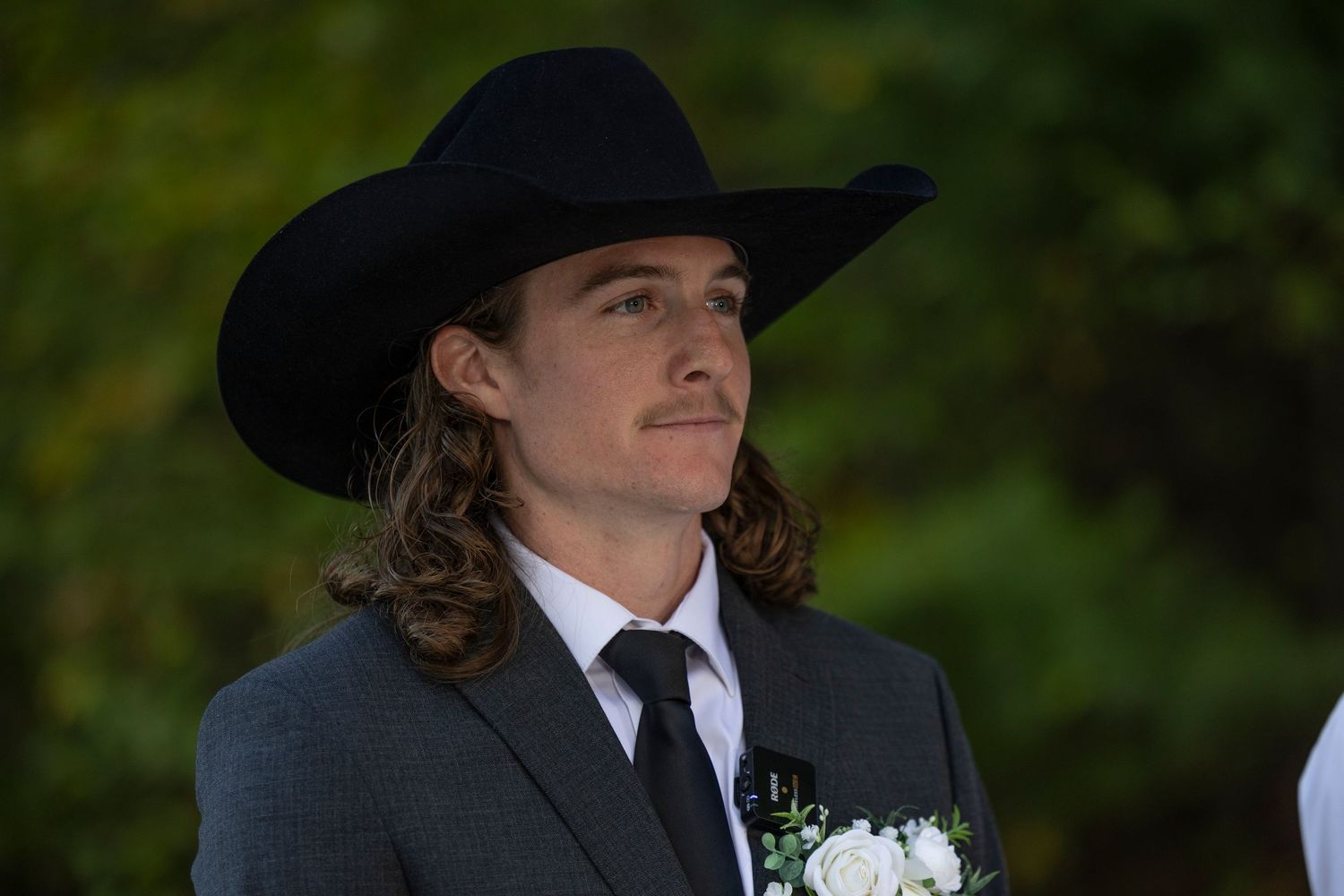 A groom in a black cowboy hat, dark suit and tie smiles while standing outdoors during a wedding ceremony.