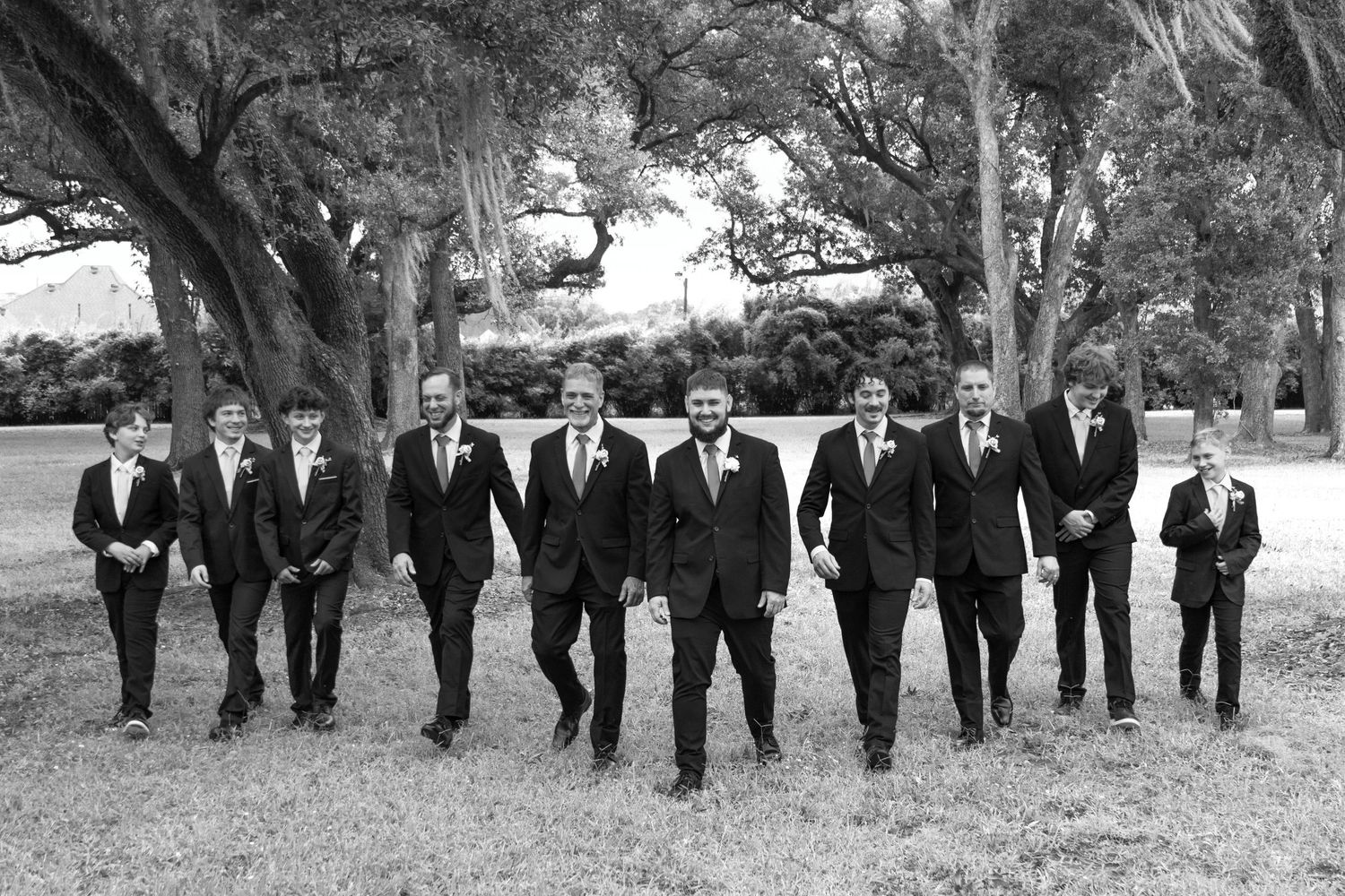 A group of people in formal black suits walk in a line across a grassy outdoor setting during a special occasion.