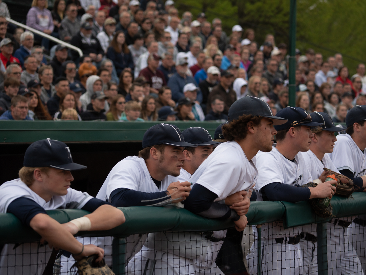 Baseball players in white uniforms lean on dugout railing watching game with large crowd in background.