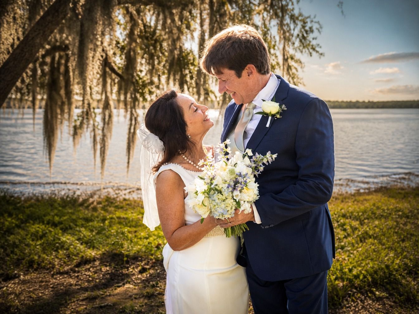 Bride and groom share a romantic moment by a lakeside, bride holding white bouquet, groom in navy suit at sunset.