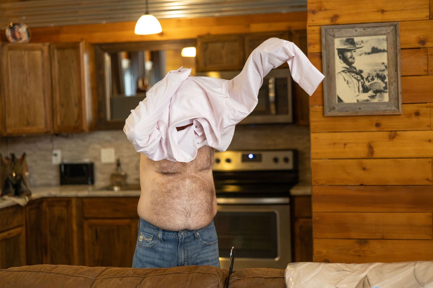 Man removing a pink shirt in a rustic wood-paneled kitchen with cabinets and stove visible in the background.