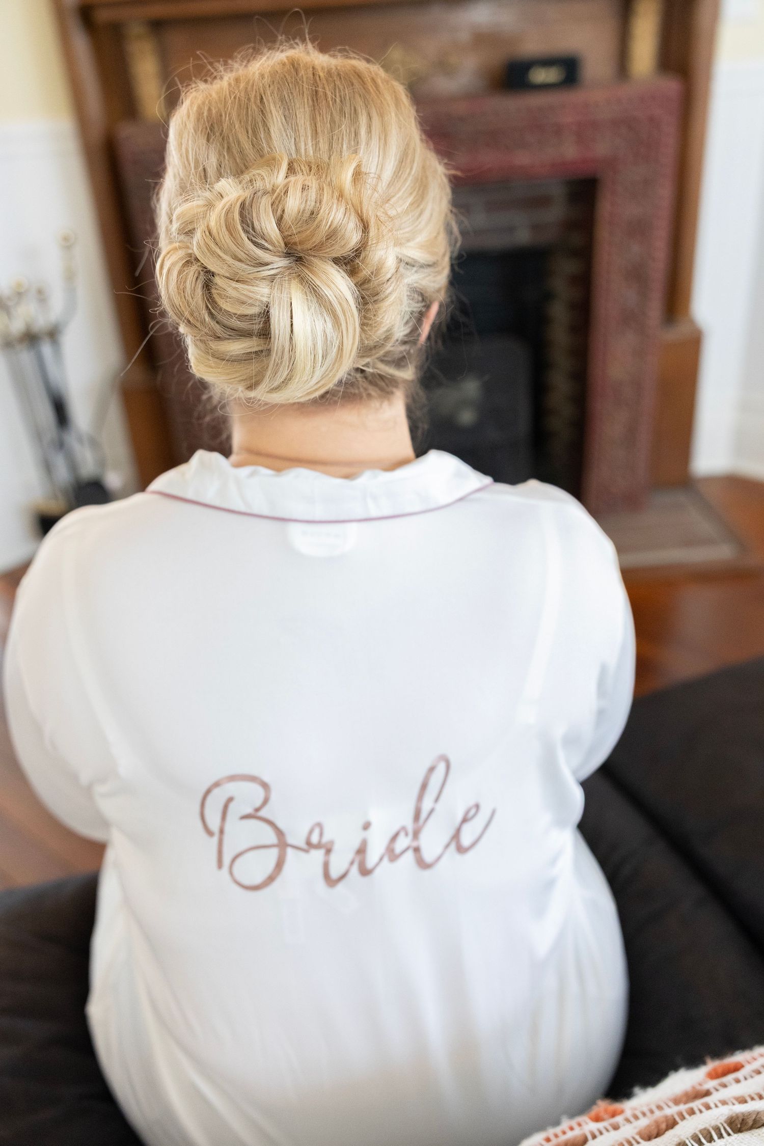 Bride wearing white satin robe with rose gold script, elegant updo hairstyle, seated before fireplace on wedding day.
