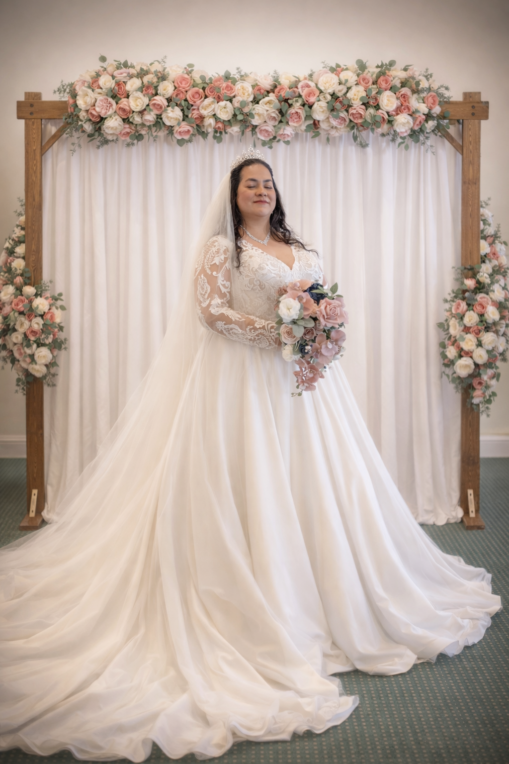 Bride in white lace ball gown with veil holding pink bouquet before floral arch.