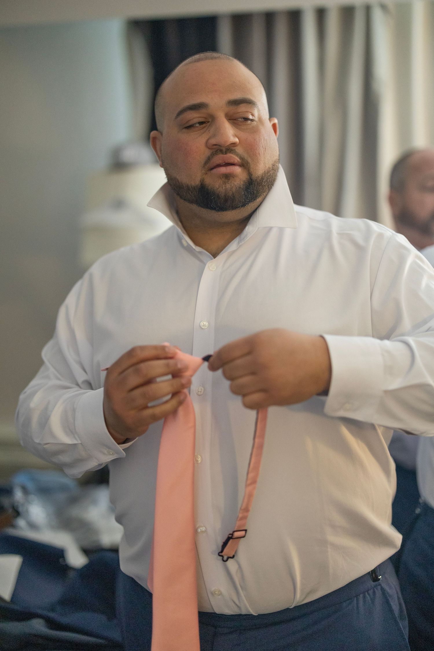A man in a white dress shirt tying a pink necktie, preparing for a formal event.