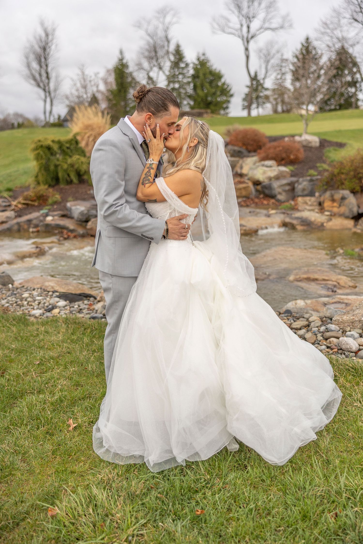 Newlyweds in formal wedding attire embrace on a grassy hill with rocks and autumn trees in the background.