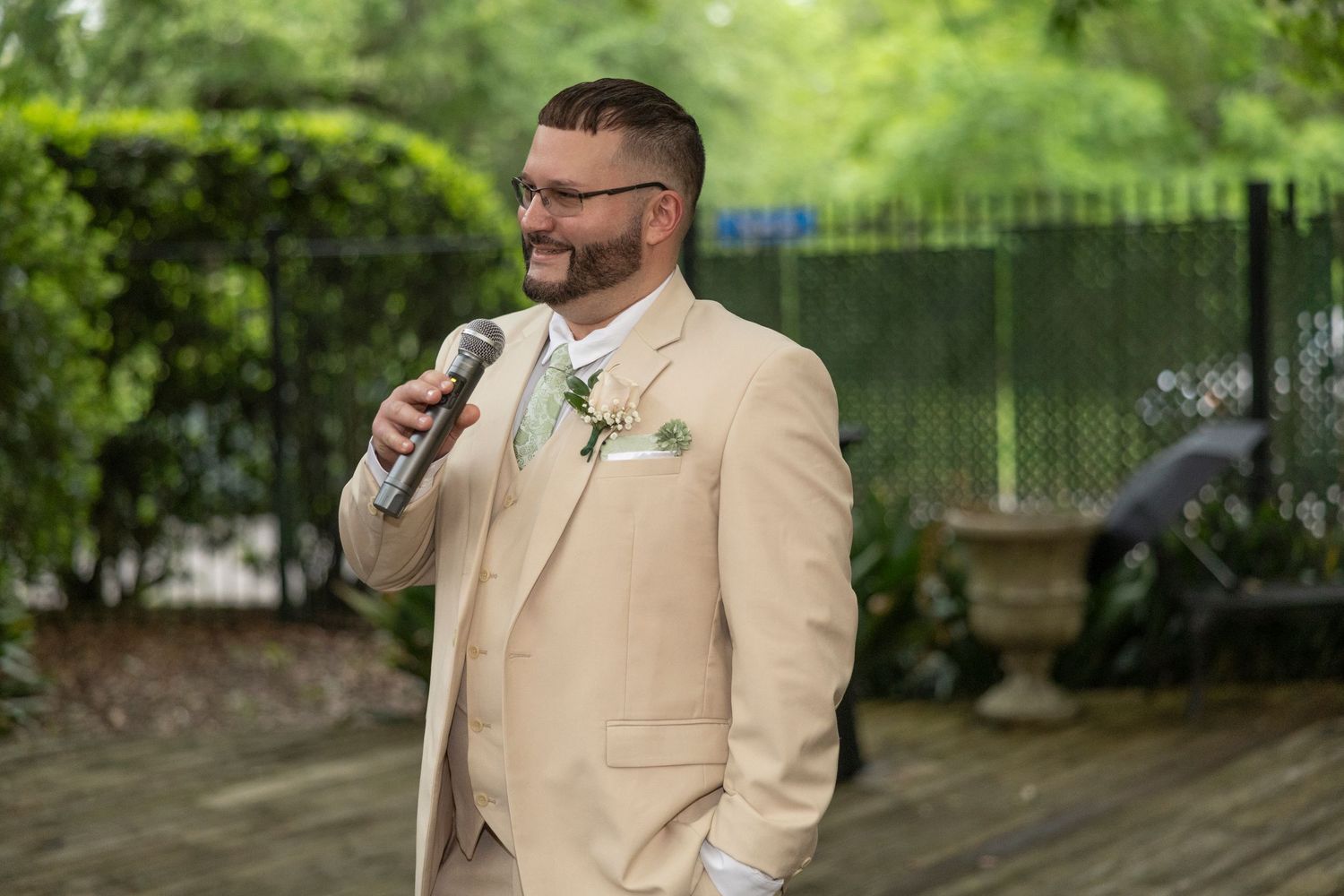 A person in a beige suit and glasses speaks into a microphone at an outdoor event with greenery in the background.