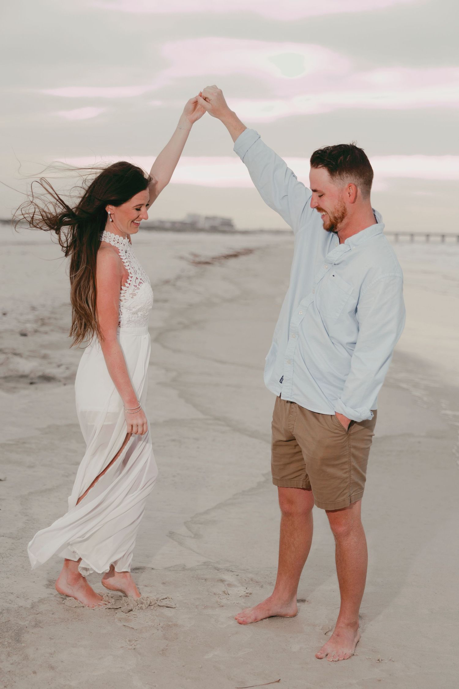 A couple dances barefoot on a sandy beach at sunset, the woman in a white dress being twirled by a man in a blue shirt.
