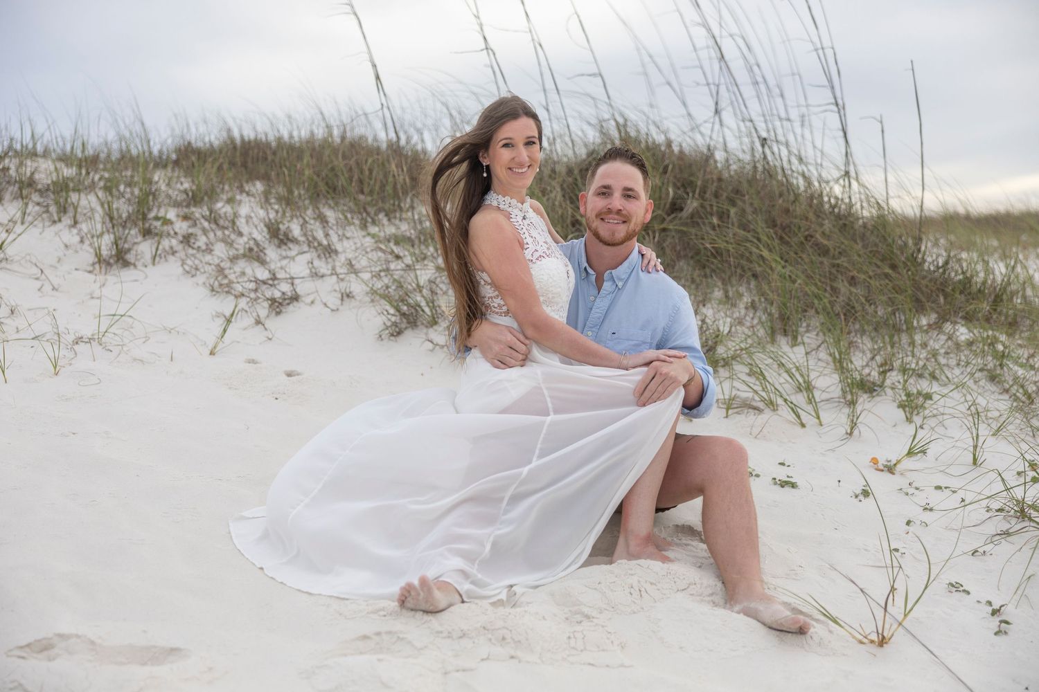 Couple posing on white sand beach dunes, woman in flowing white dress, man in light blue shirt, smiling together.