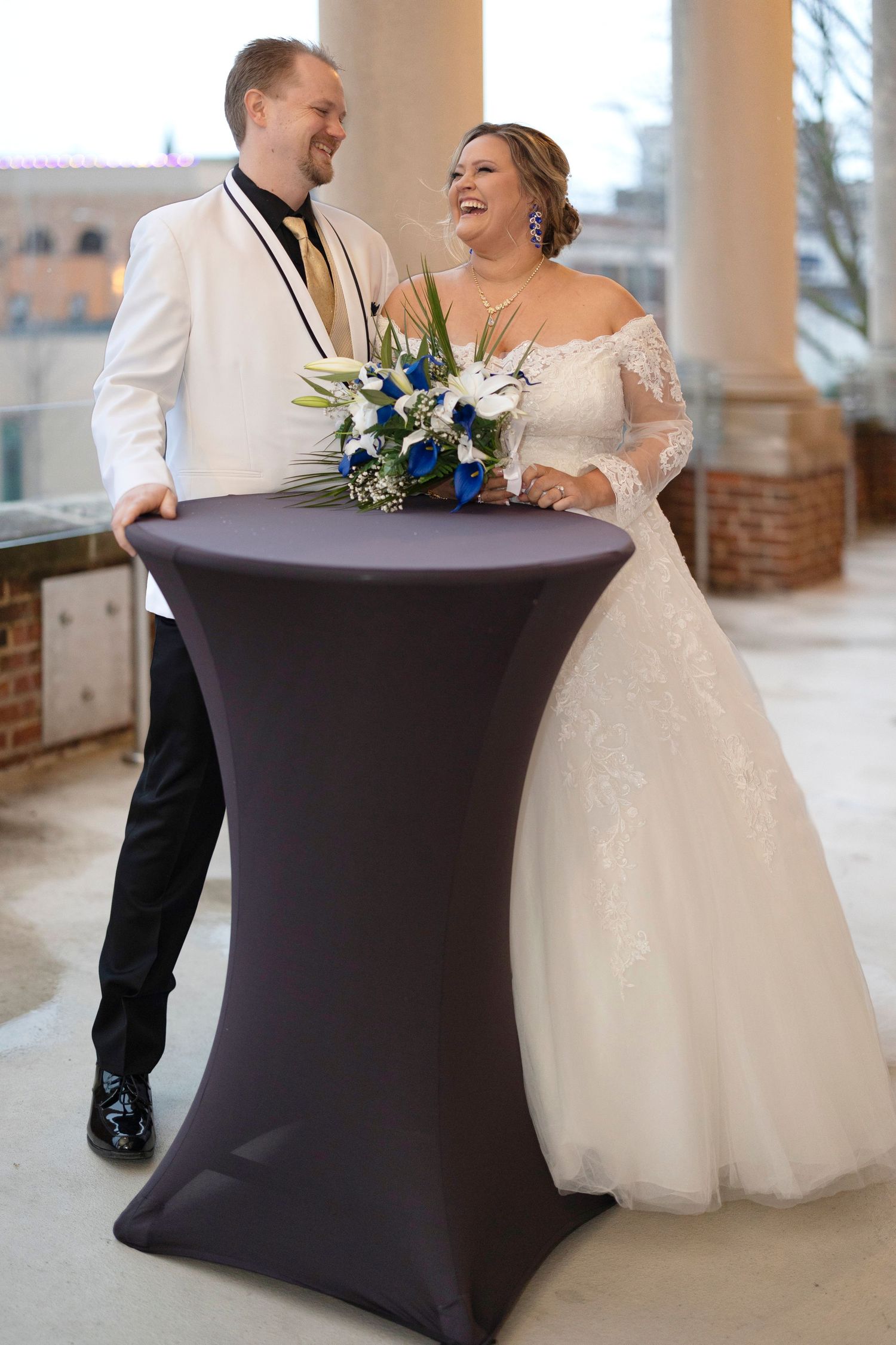 Smiling bride and groom standing at a cocktail table outdoors, bride holding blue and white bouquet in wedding attire.