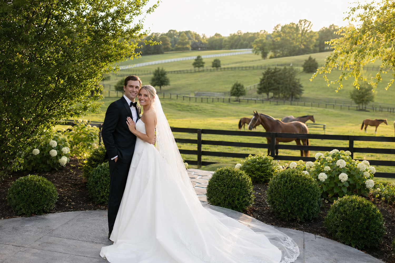 Bride and groom pose on a patio with lush green fields, white hydrangeas, and horses by a fence at golden hour.