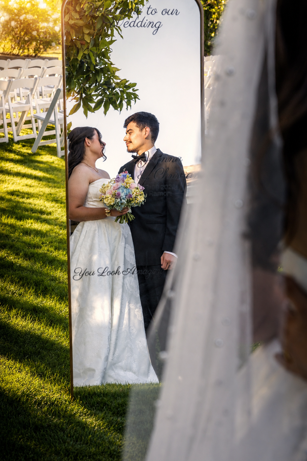 Bride and groom share a romantic moment at outdoor wedding ceremony, reflected in a decorative mirror on green lawn.