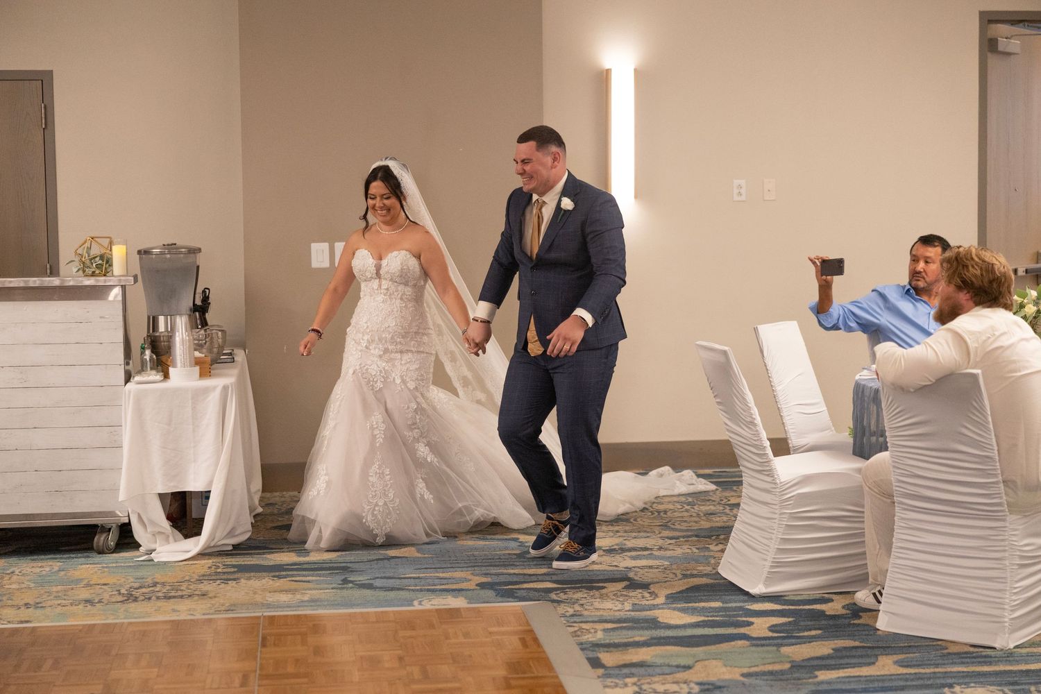 Bride in white gown and groom in navy suit enter wedding reception hall, greeted by smiling guests.