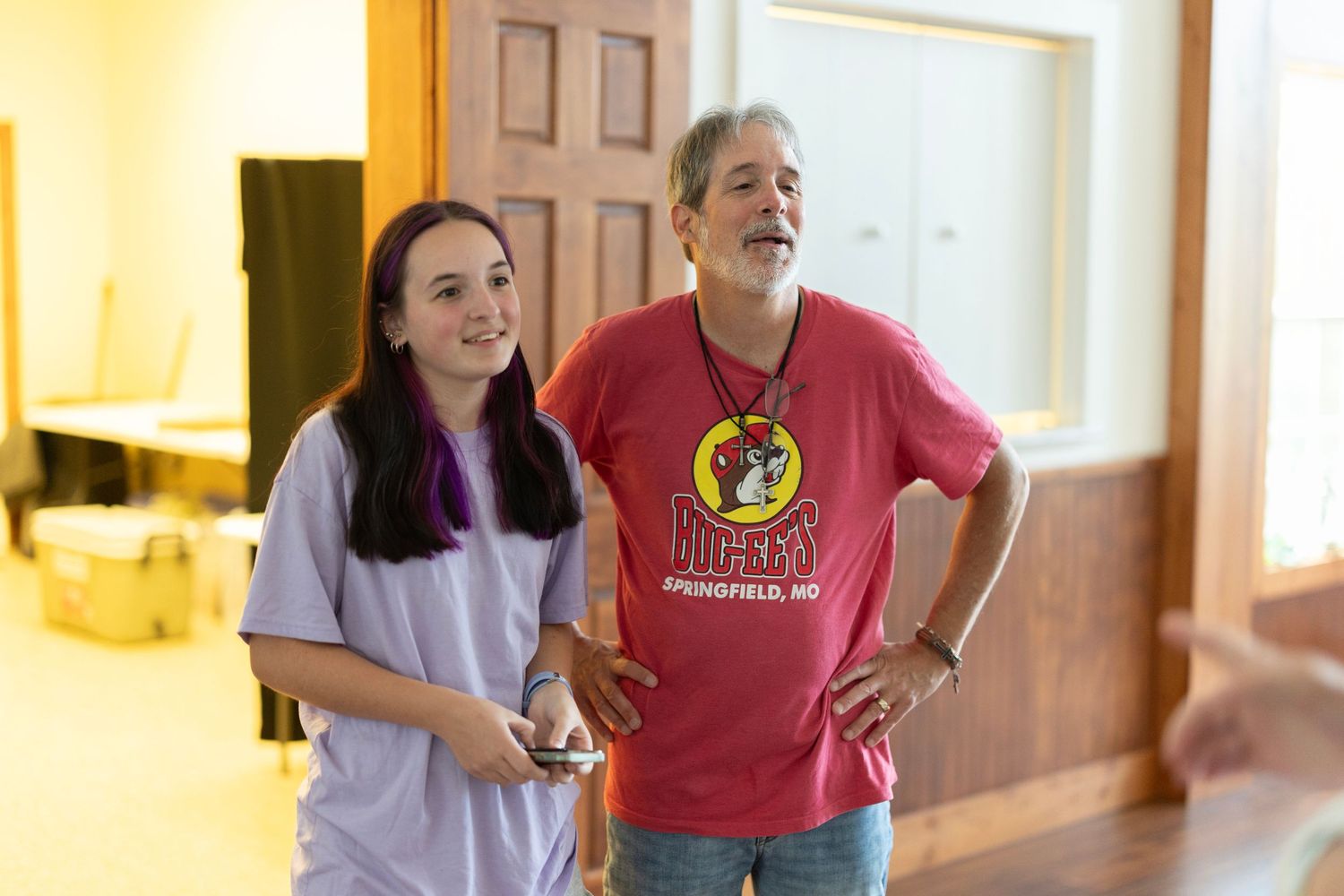 A young girl and an older man smiling together indoors, man wearing a red Buc-ee's Springfield, MO t-shirt.