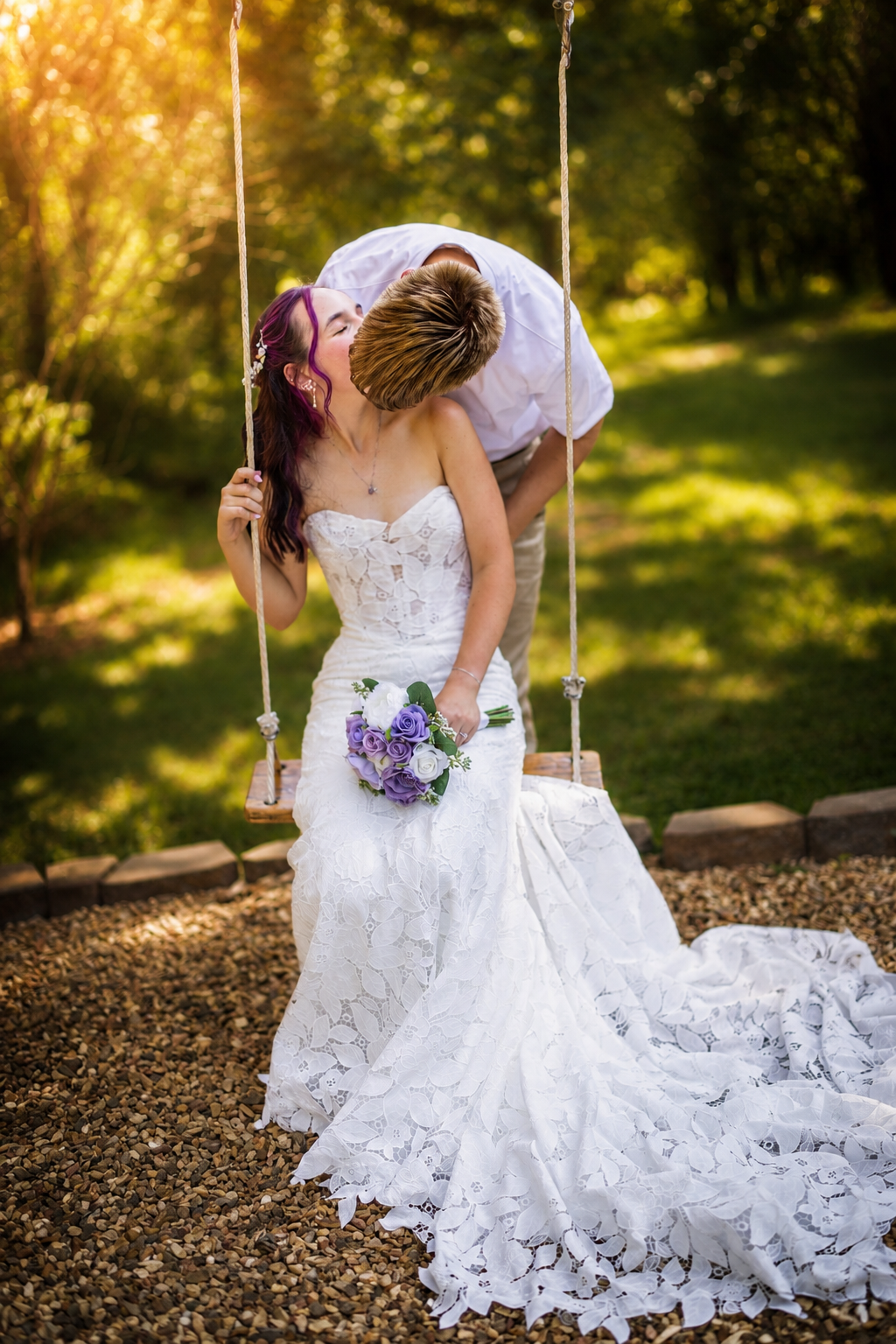Groom leans down to kiss bride seated on rustic rope swing in lace wedding gown during golden hour outdoors.