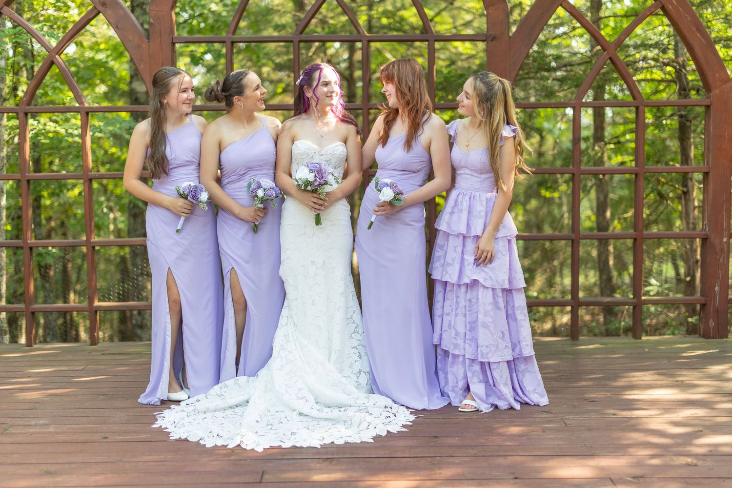 Bride in white lace gown poses with four bridesmaids in lavender dresses, holding bouquets in a wooden garden gazebo.