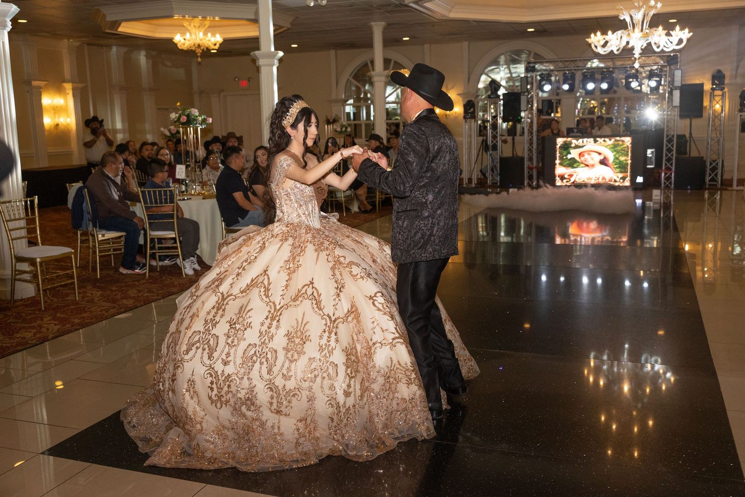 A young woman in a gold ballgown dances with a partner in a black hat at an elegant quinceañera celebration.