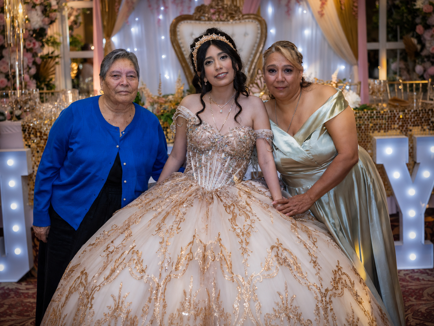 Young woman in gold quinceañera gown poses with two women at elegant celebration party.