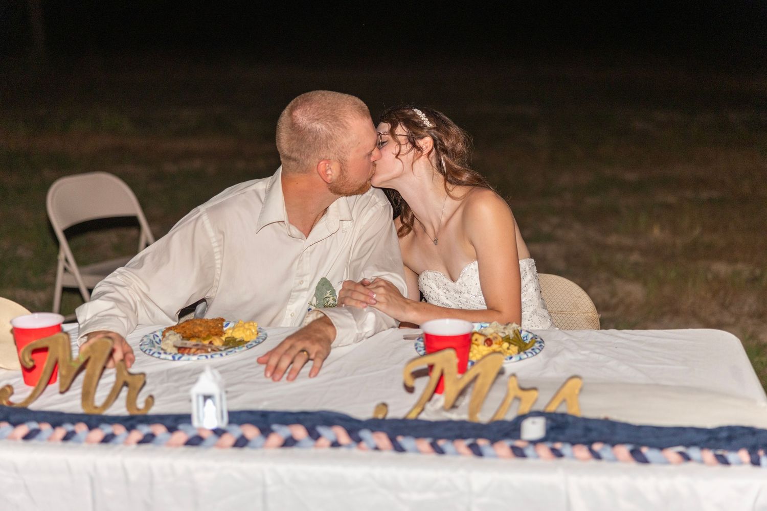 Newlywed couple sharing a kiss at their outdoor wedding reception sweetheart table decorated with Mr. and Mrs. signs.