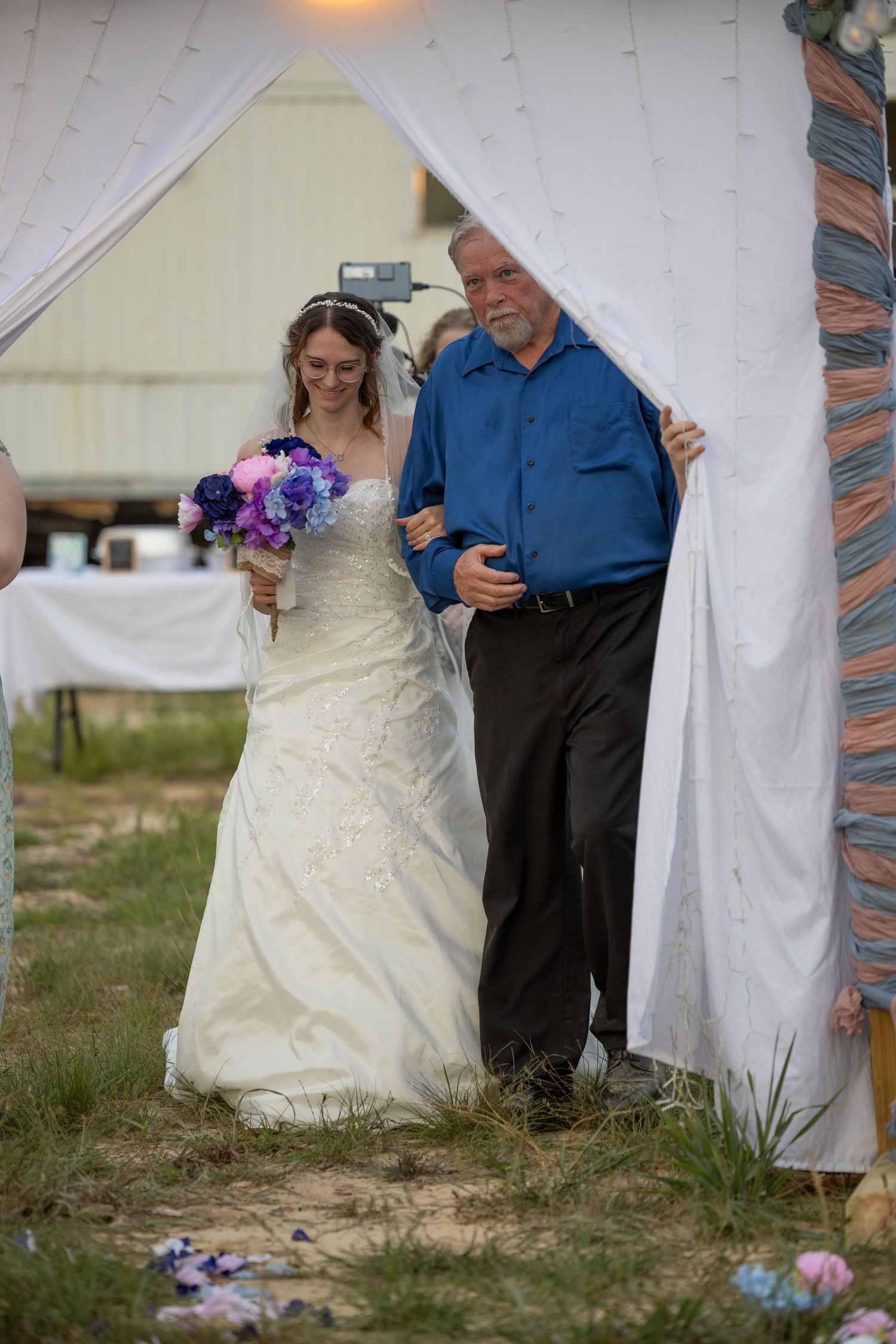 Bride in white gown holding purple bouquet walks with older man in blue shirt through draped wedding arch.