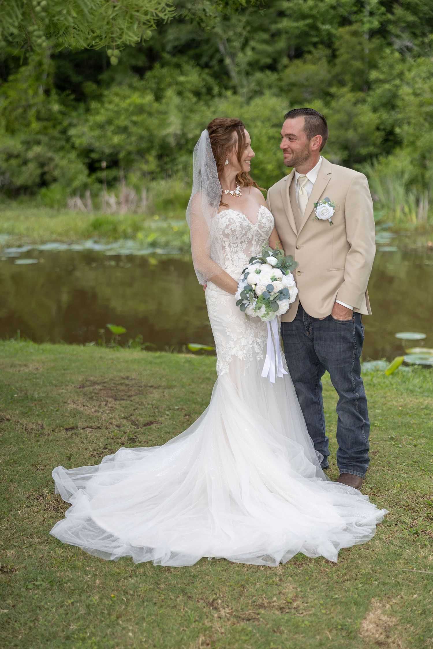 Newlyweds pose together by a scenic pond surrounded by lush greenery in their wedding attire.