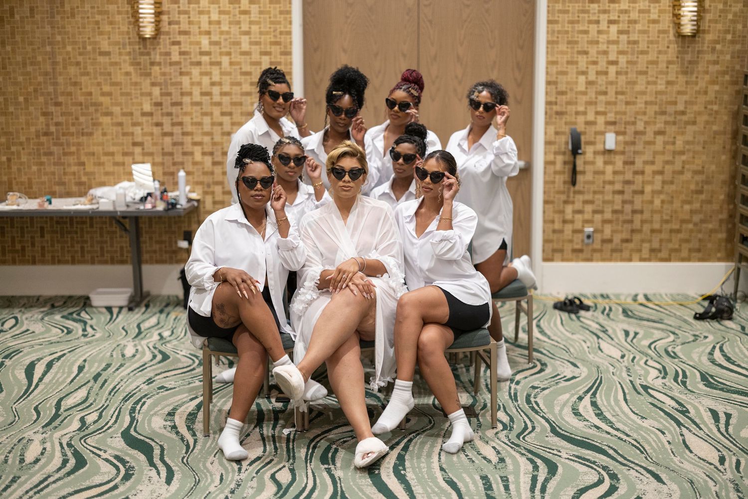 A group poses in matching white robes and sunglasses in front of a beige brick wall.