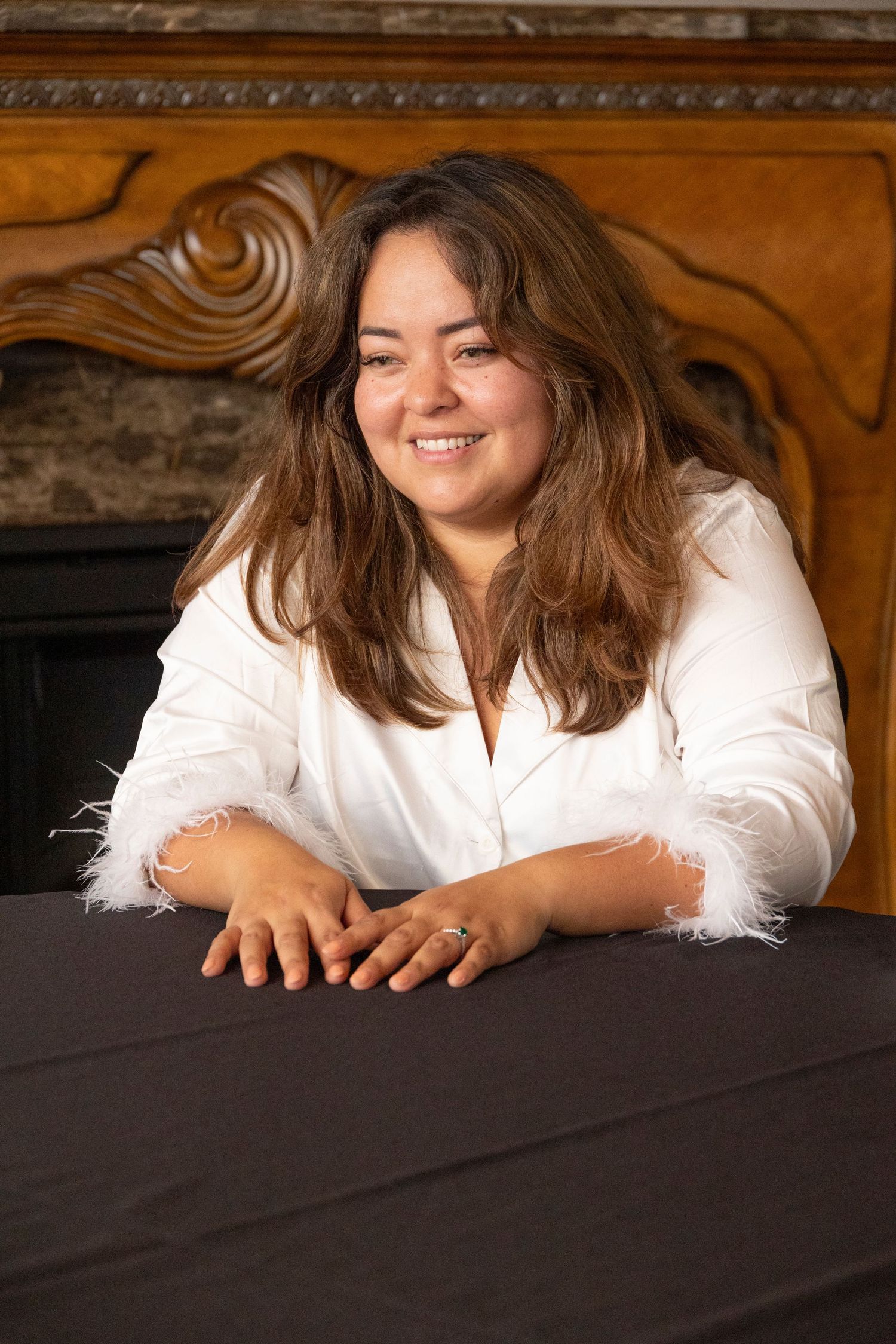 Smiling person in white blouse leaning on dark table with ornate wooden headboard in background.