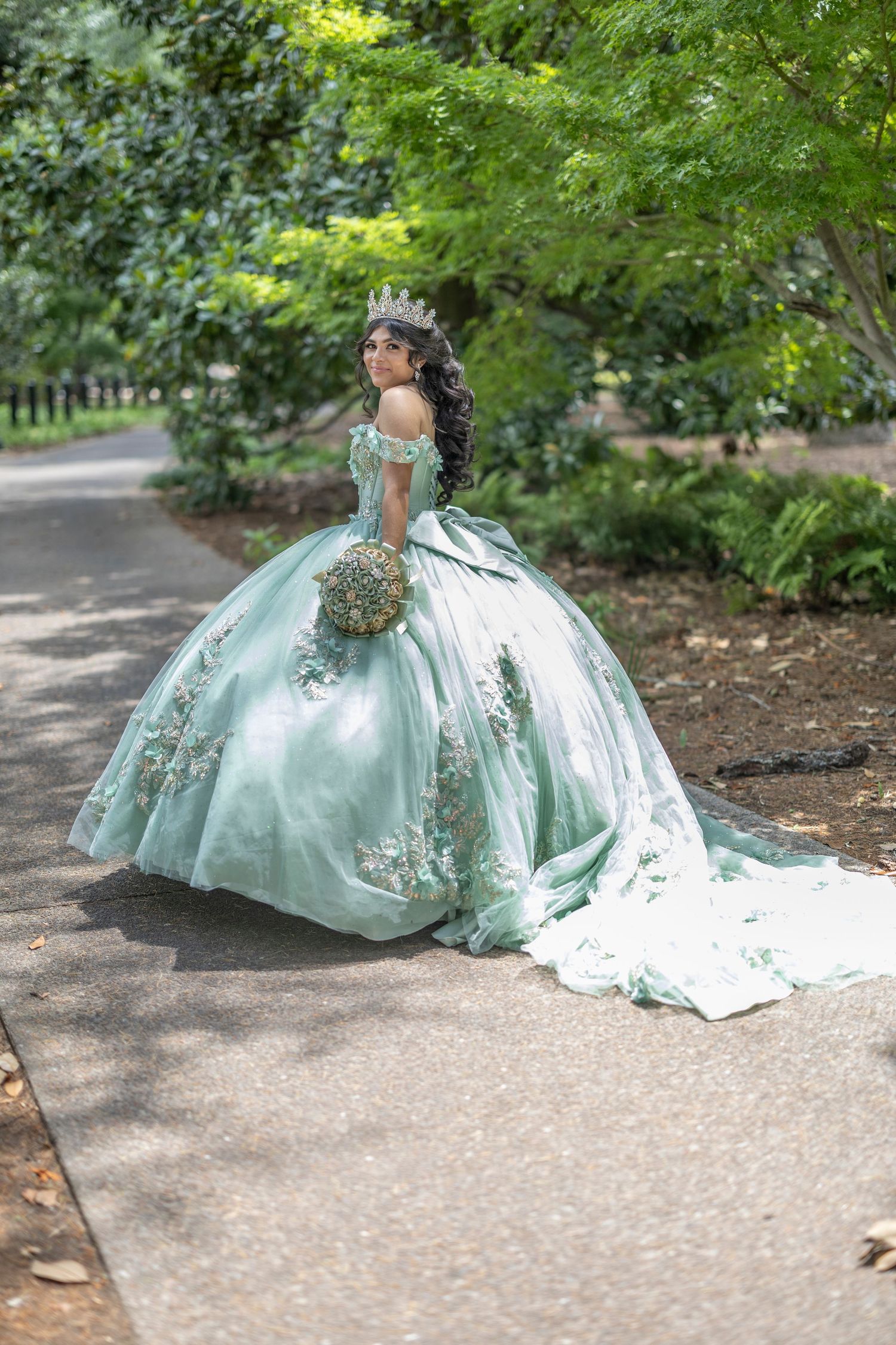 A stunning mint green quinceañera ball gown with ornate embellishments and long train photographed in a garden path setting.