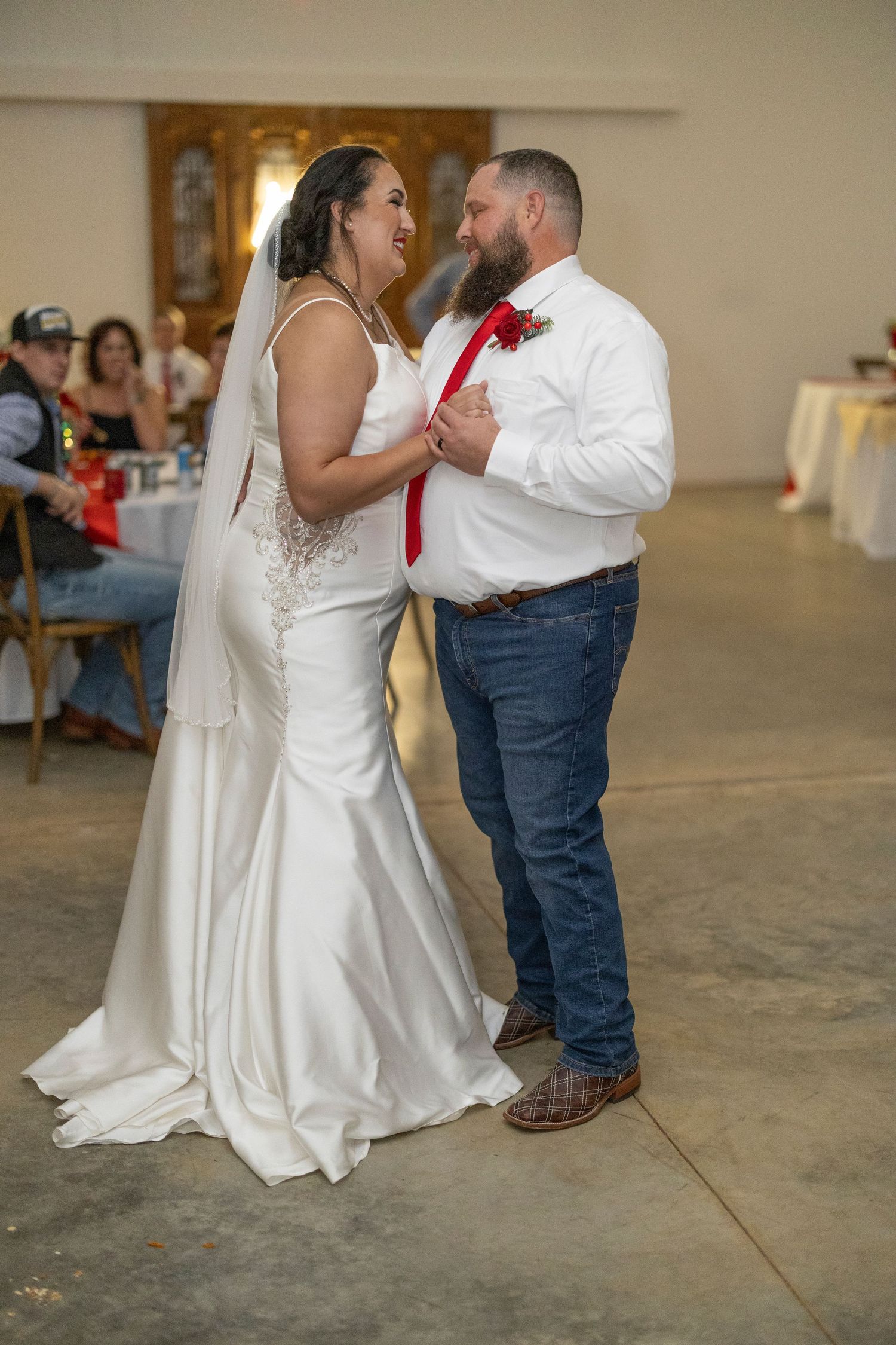 A couple shares their first dance at a wedding reception, the bride in a fitted white gown and groom in jeans and red tie.