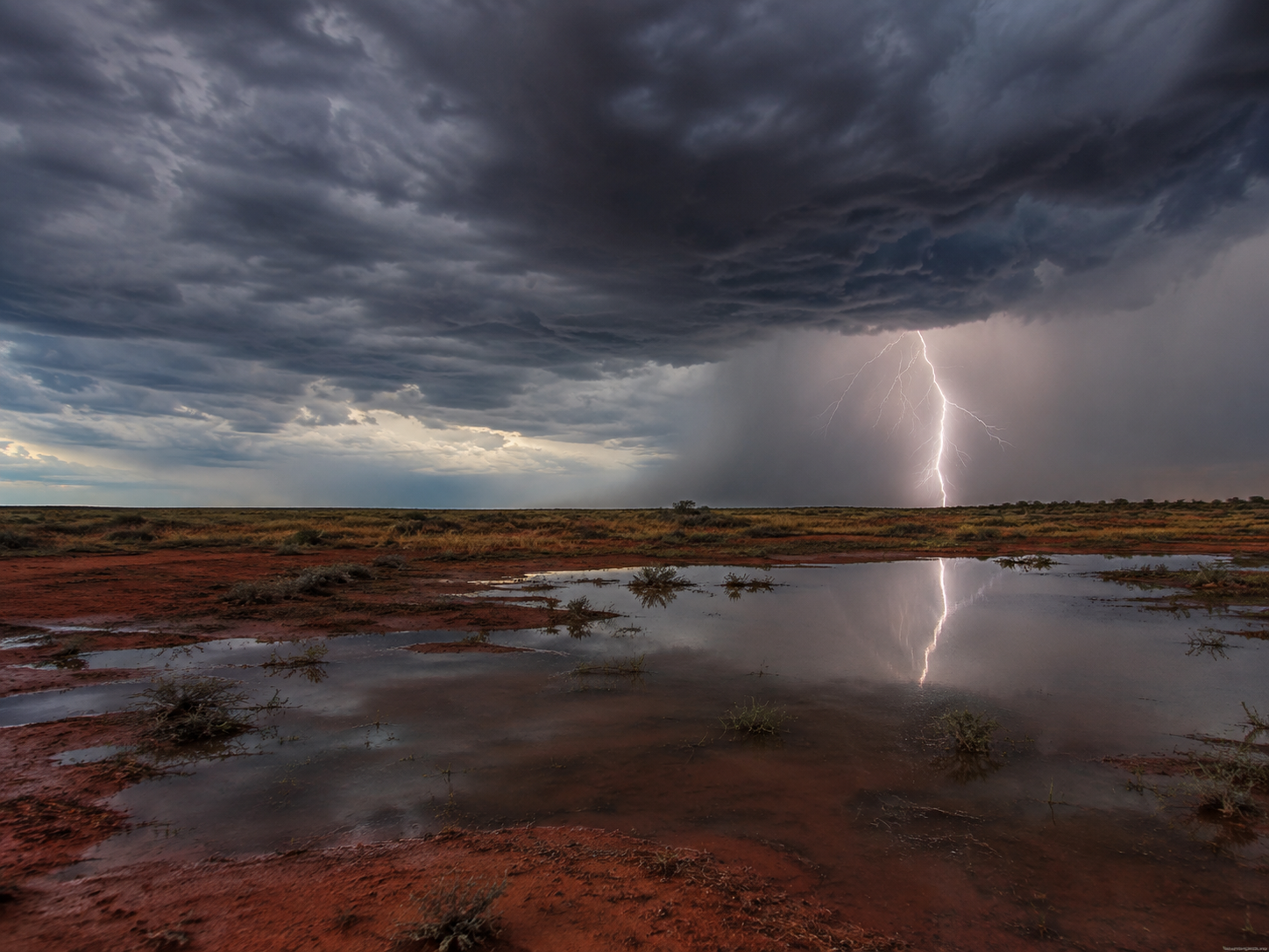 Lightning strikes over a red desert landscape with dramatic storm clouds reflected in a shallow puddle.