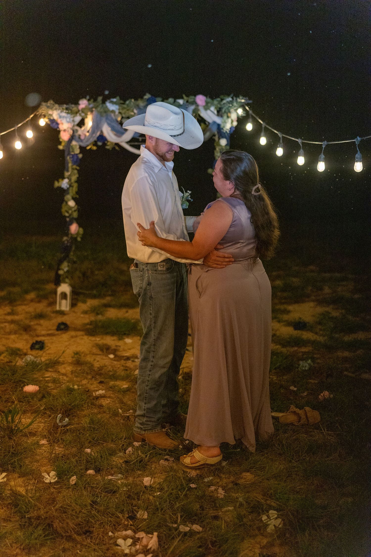 Couple sharing a romantic dance outdoors at night under string lights and floral arch, man wearing cowboy hat.