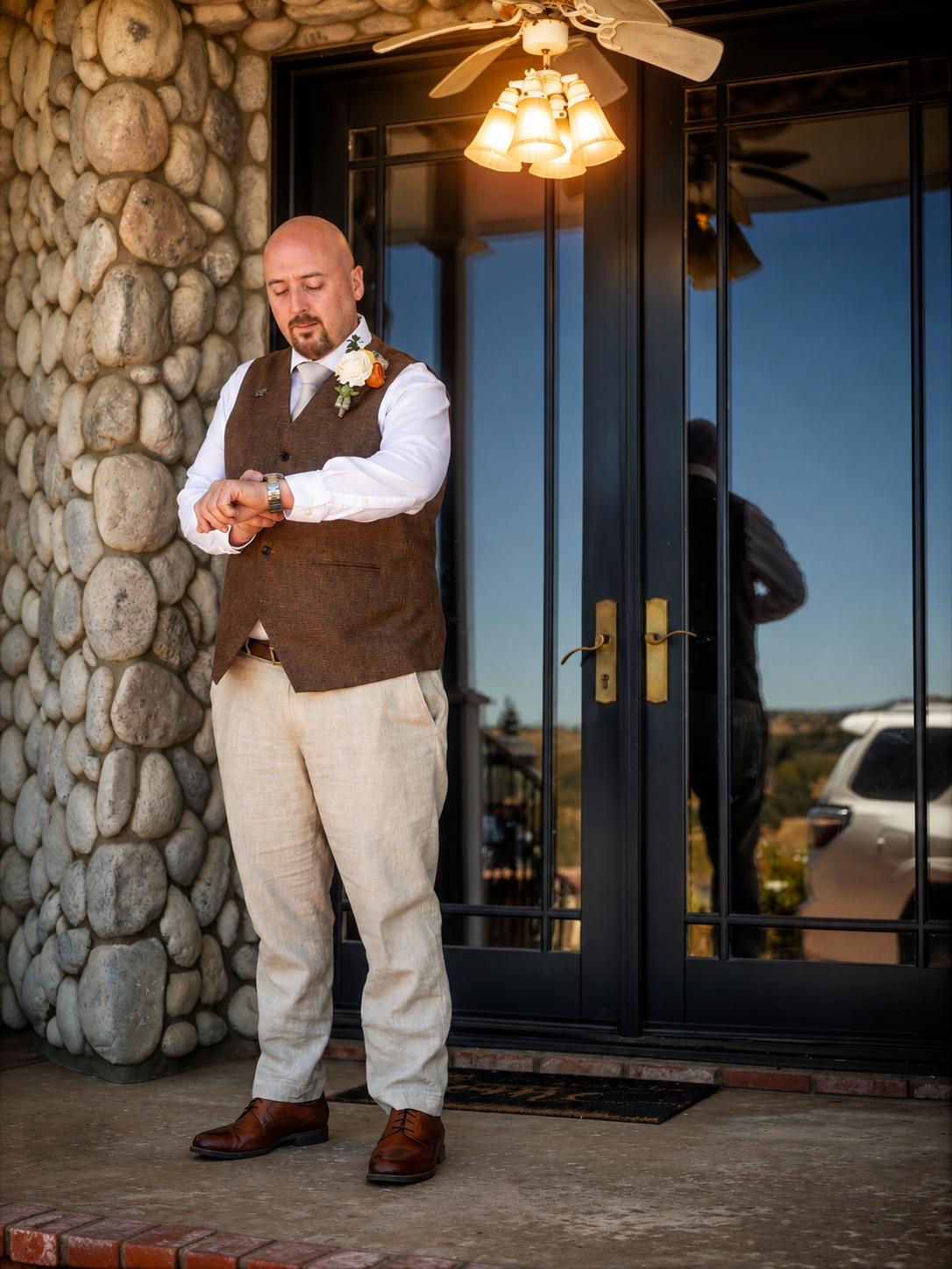 Groom in brown vest and linen pants checks his watch outside a stone building before his wedding ceremony.