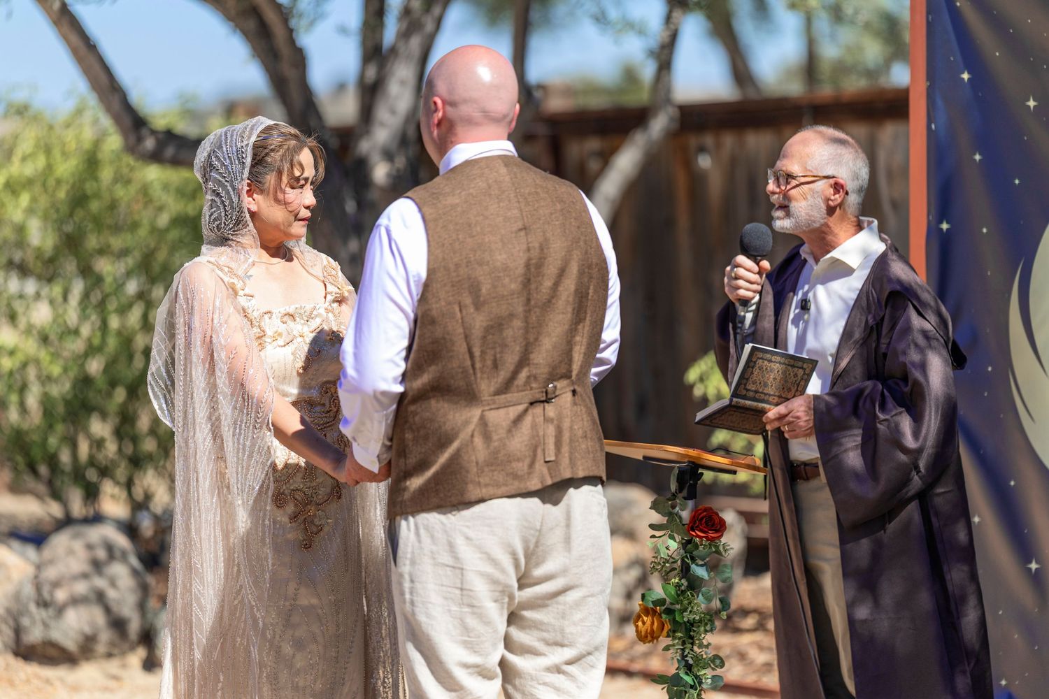 Outdoor wedding ceremony with bride in lace veil, groom in tan suit, and officiant reading vows under desert backdrop.