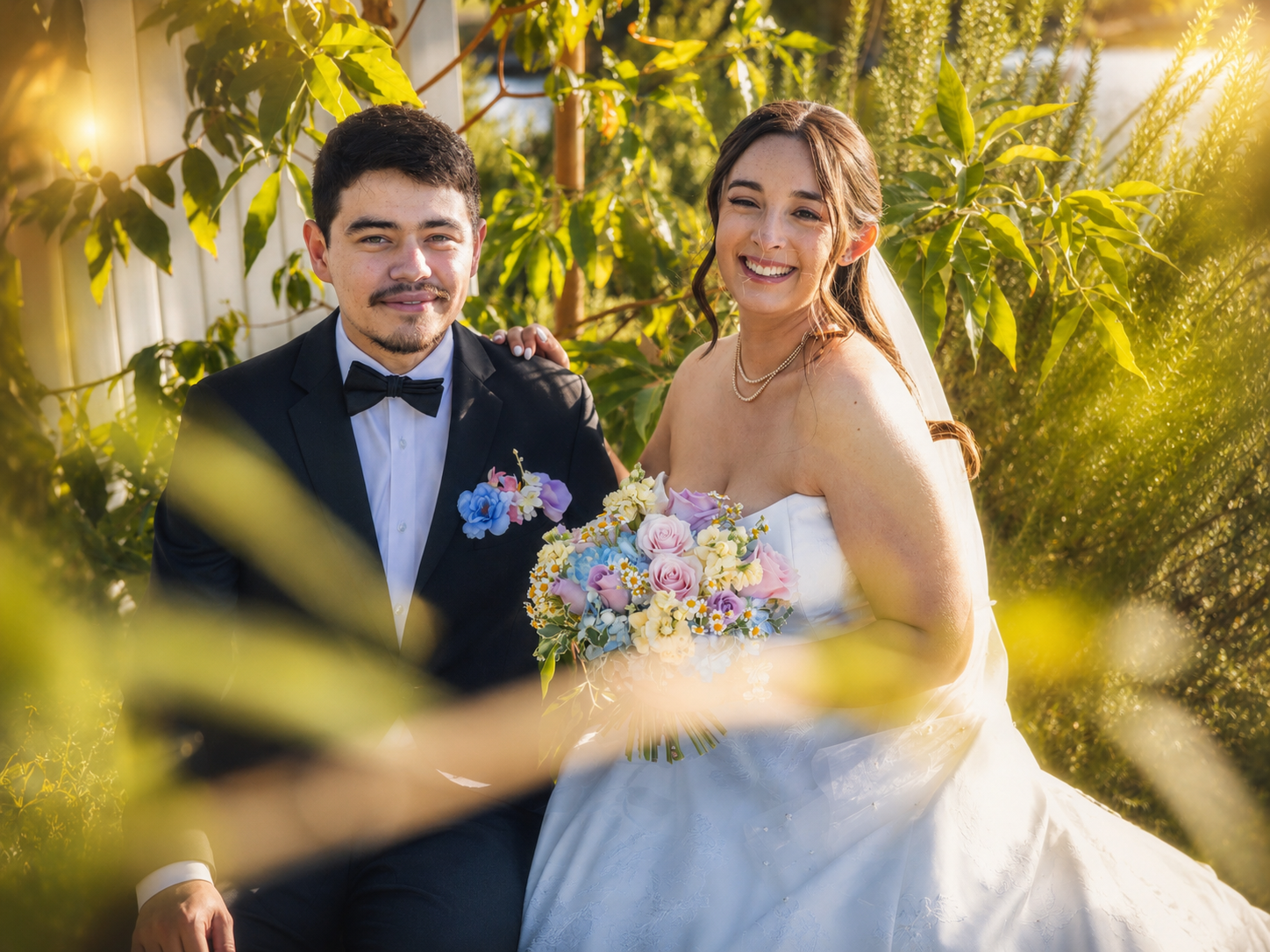 Smiling newlyweds pose outdoors in golden sunlight, groom in tuxedo, bride in white gown holding colorful bouquet.