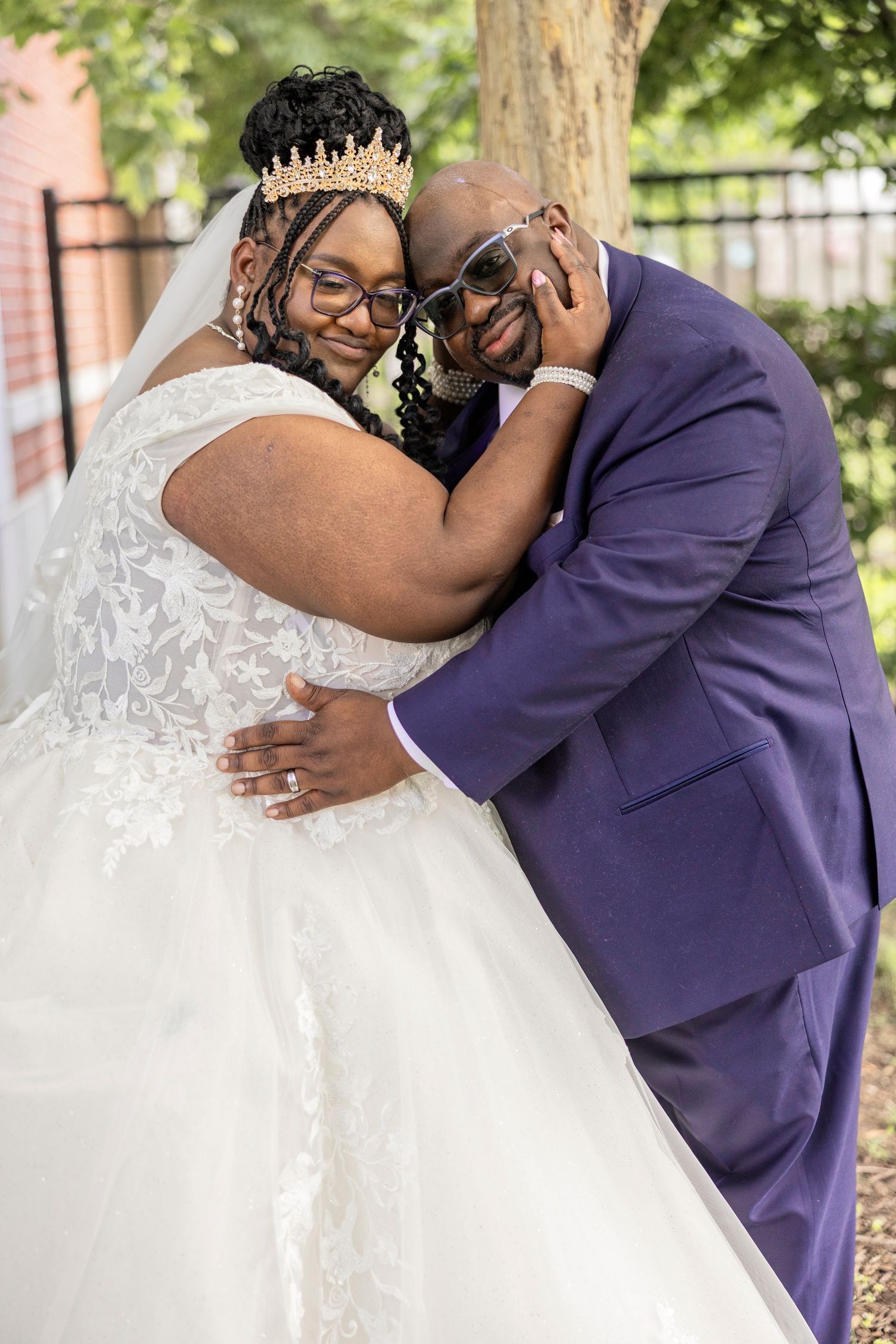 A bride in a white ballgown and crown embraces her groom in a purple suit outdoors on their wedding day.