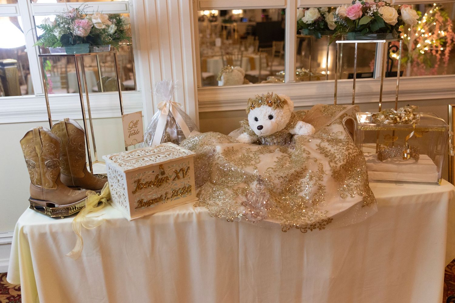 Gold quinceañera gift table with cowboy boots, decorated box, sparkly clutch, and teddy bear at elegant venue.