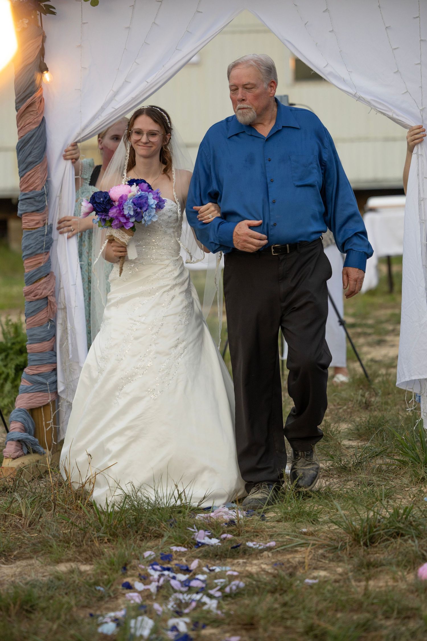 Bride in white gown holding purple bouquet walks down aisle with older man in blue shirt at outdoor wedding.