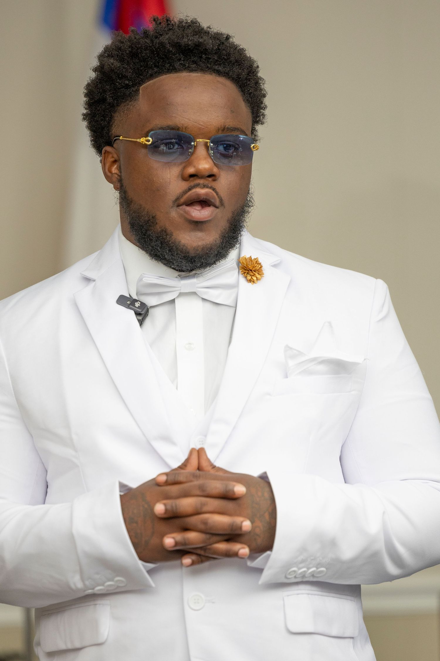 Stylish man in white suit with bow tie, gold brooch, and blue tinted sunglasses, hands clasped together.