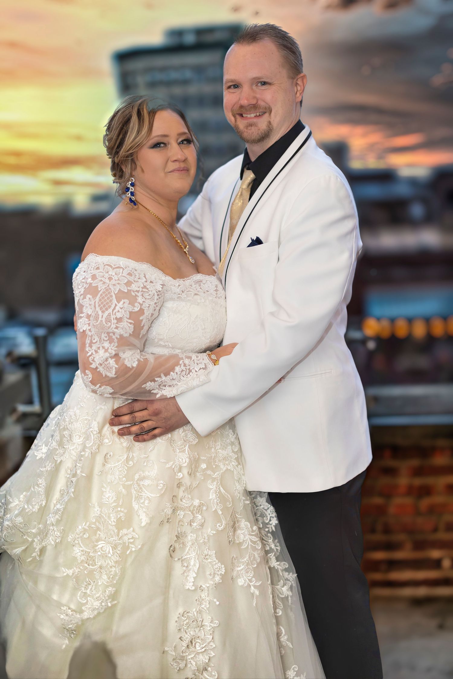 Bride in lace off-shoulder gown and groom in white tuxedo pose together at sunset rooftop wedding photo.