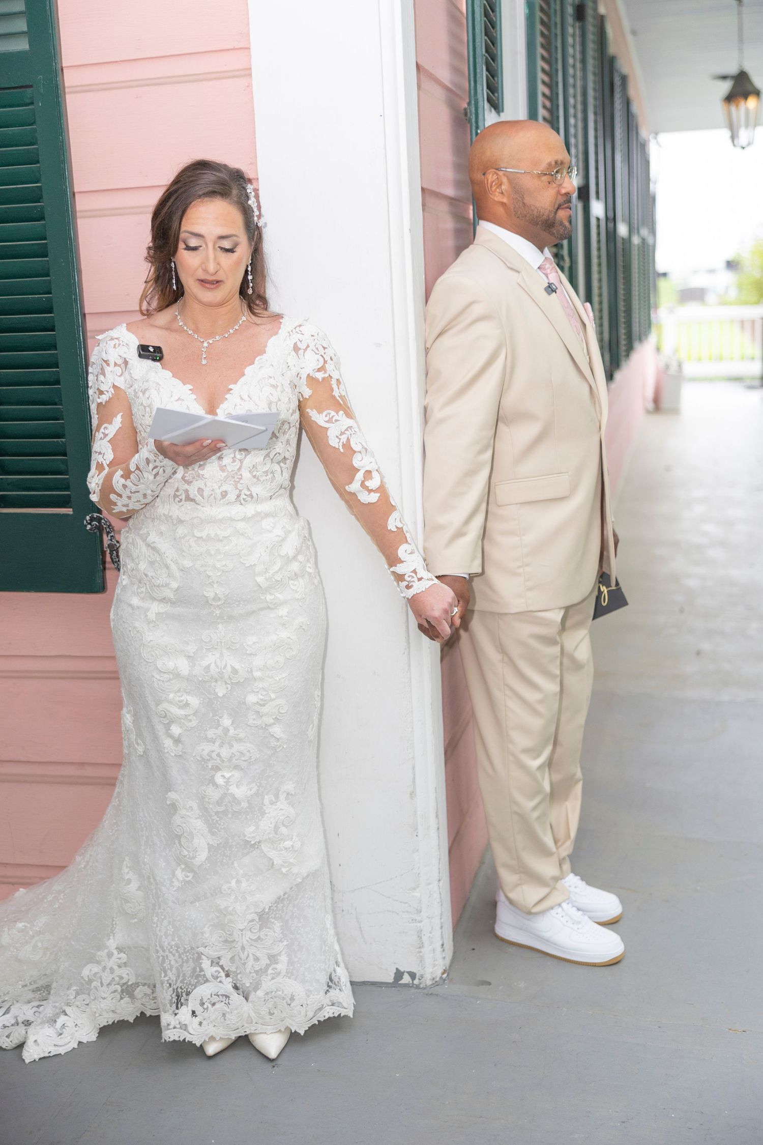 Wedding couple in formal attire walk together along a covered porch with pink walls and green shutters.