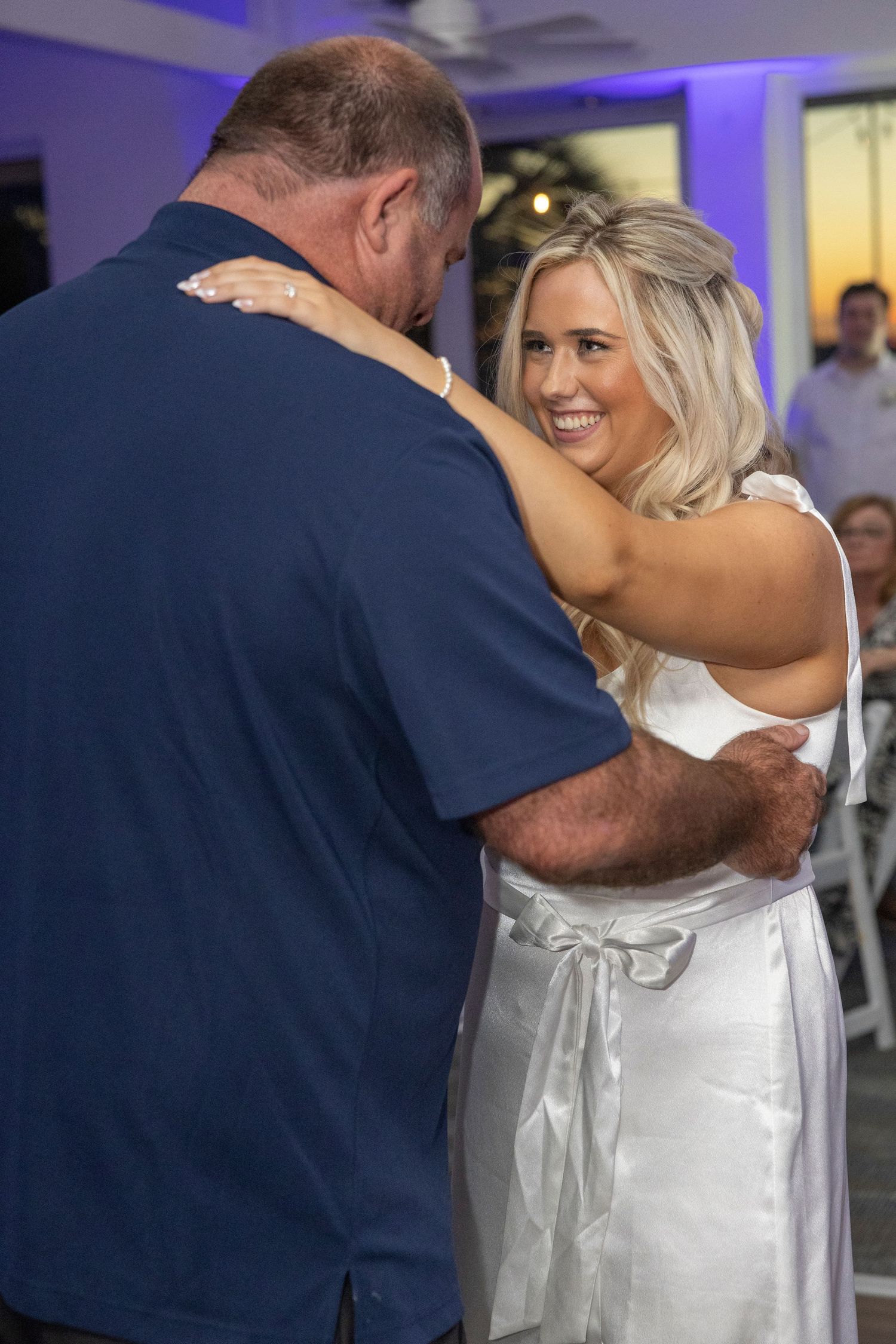 A bride in a white satin dress shares a dance with someone wearing a navy blue shirt at a wedding reception.