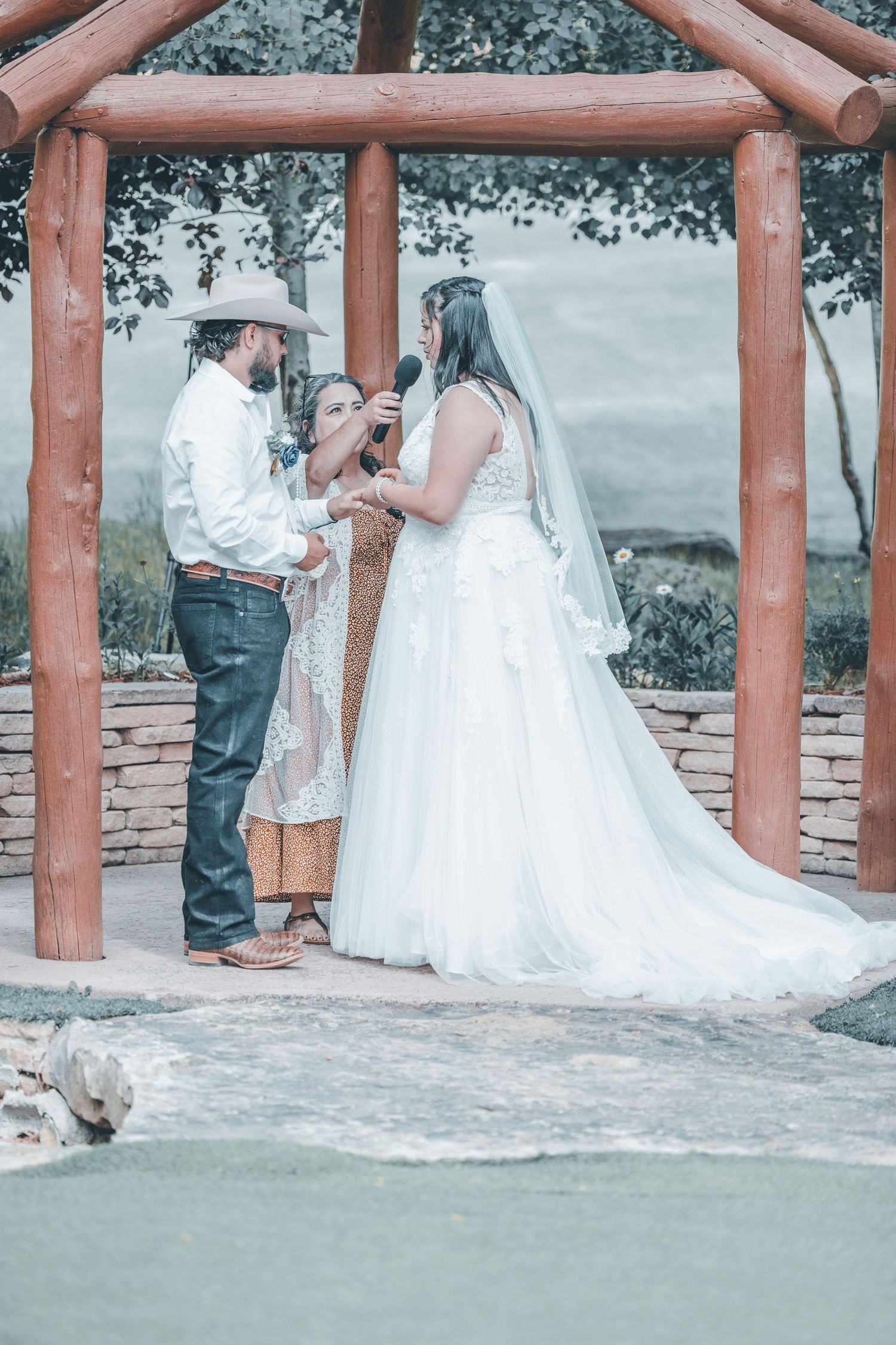 A wedding ceremony takes place under a rustic wooden pergola with stone pillars on a beautiful outdoor setting.