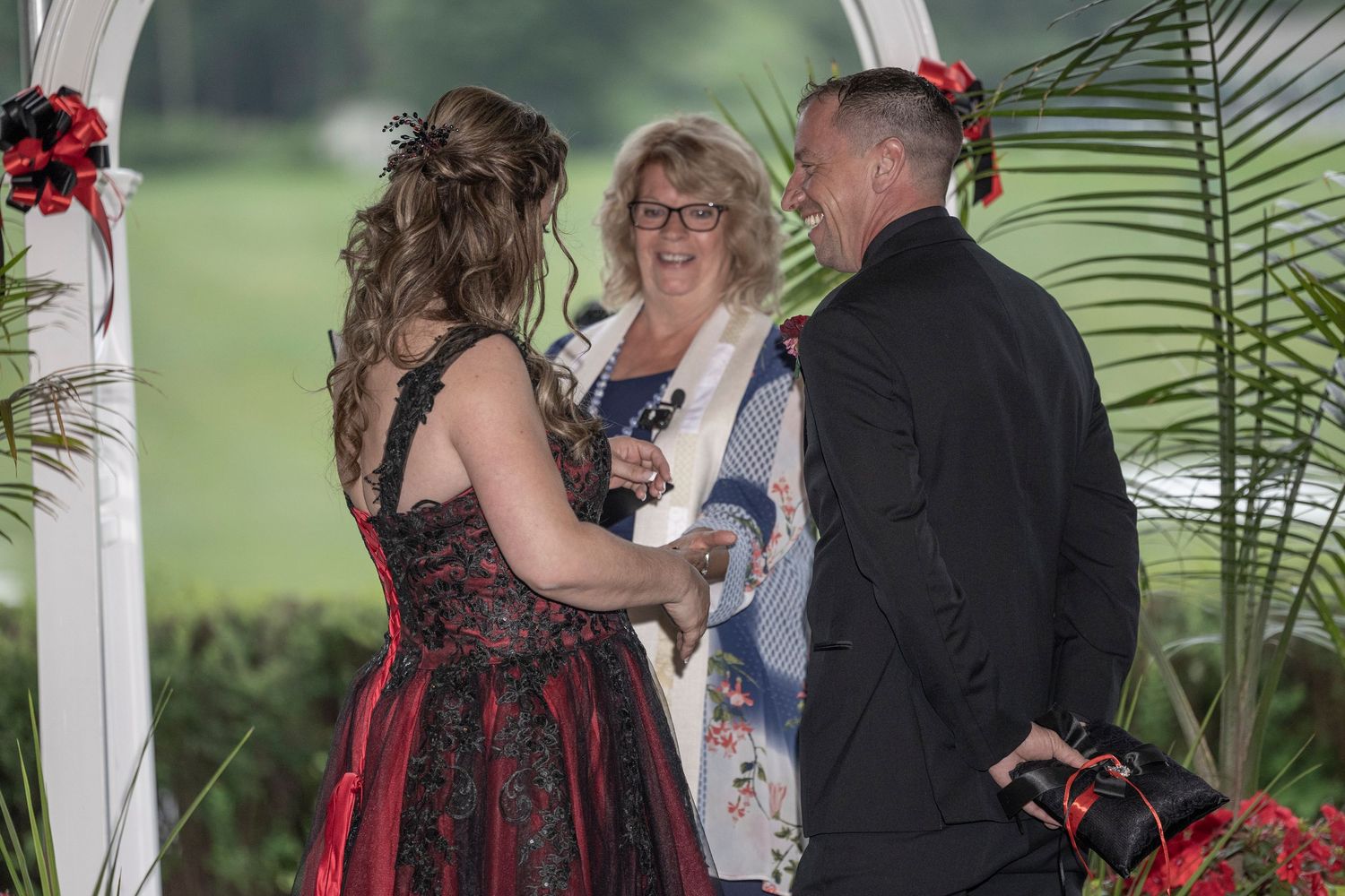 Wedding ceremony takes place under a white gazebo decorated with red flowers and tropical palm leaves.