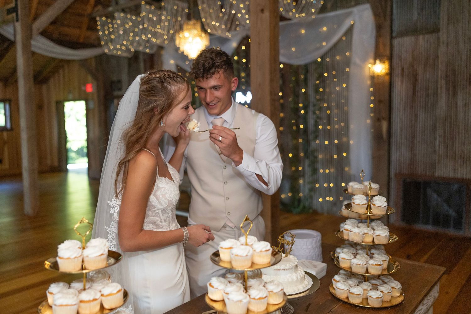 Wedding couple cuts their multi-tiered white cupcake tower display at rustic indoor reception venue with string lights.