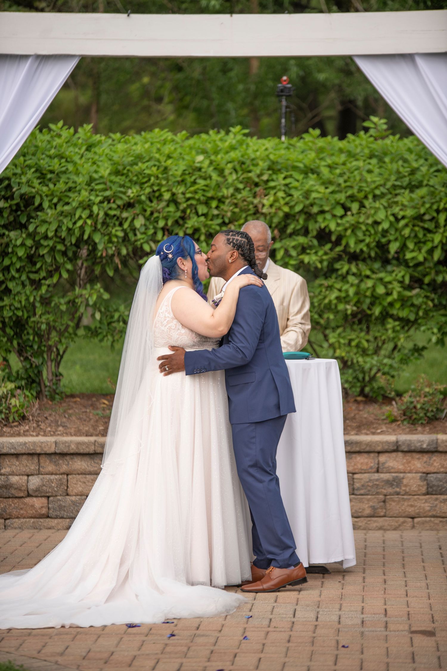 A couple in wedding attire share a romantic kiss on a brick patio surrounded by greenery and white decor.