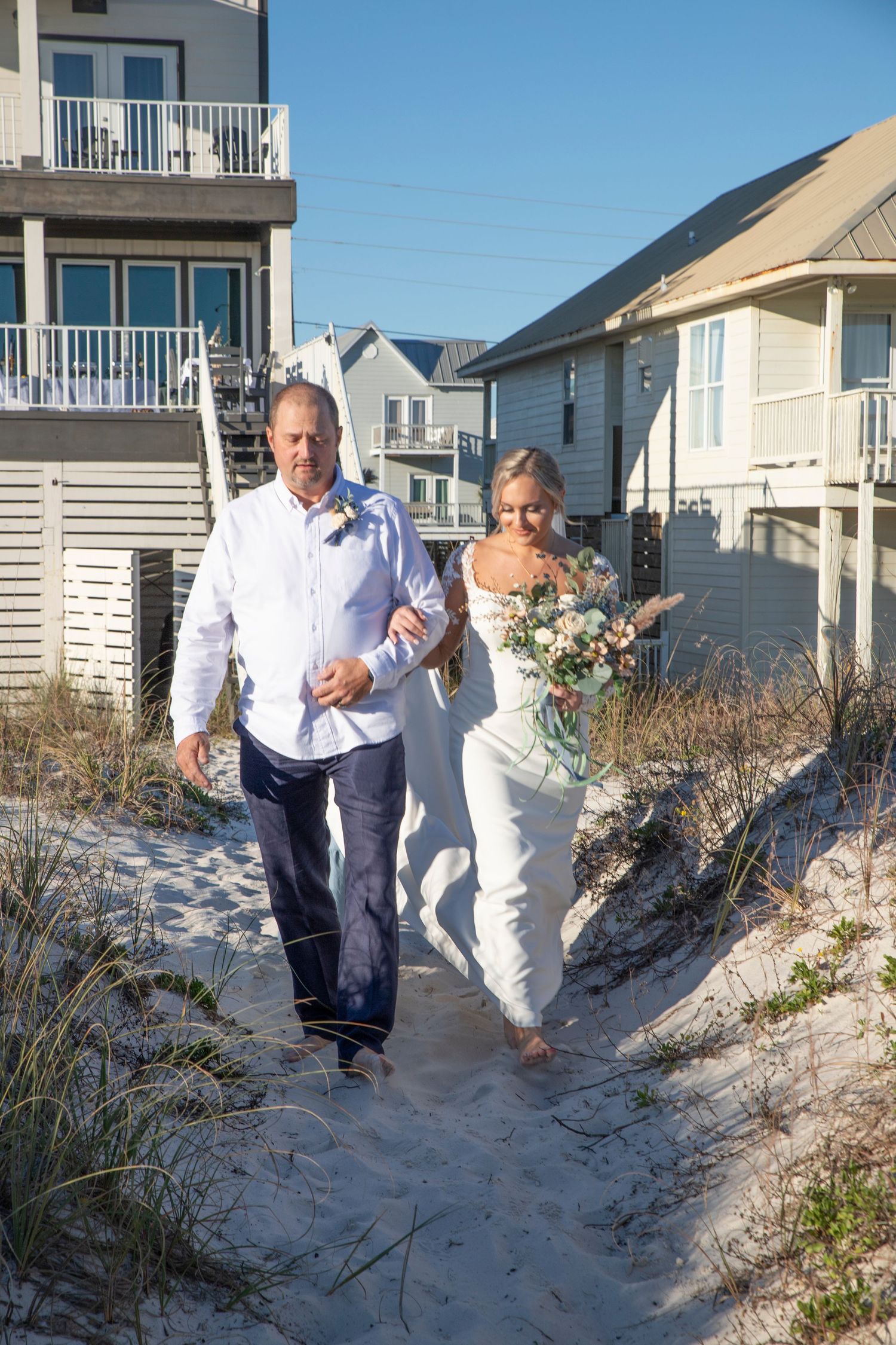 People walking along a sandy beach path between beach houses on a sunny day.
