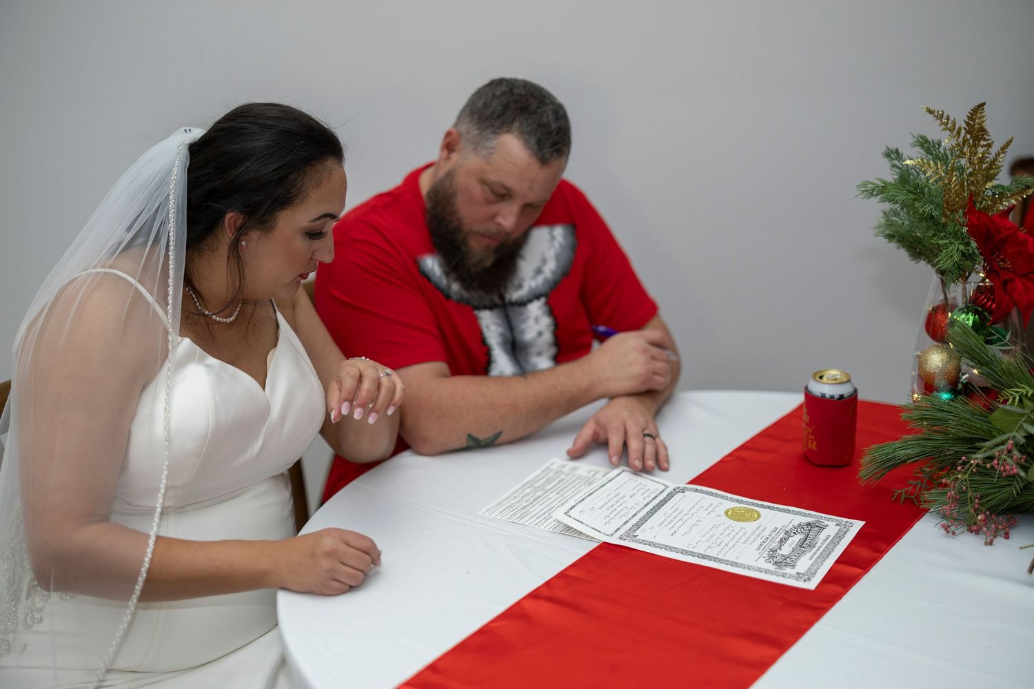 A couple in formal wedding attire signs documents at a table decorated with red tablecloth and Christmas decorations.