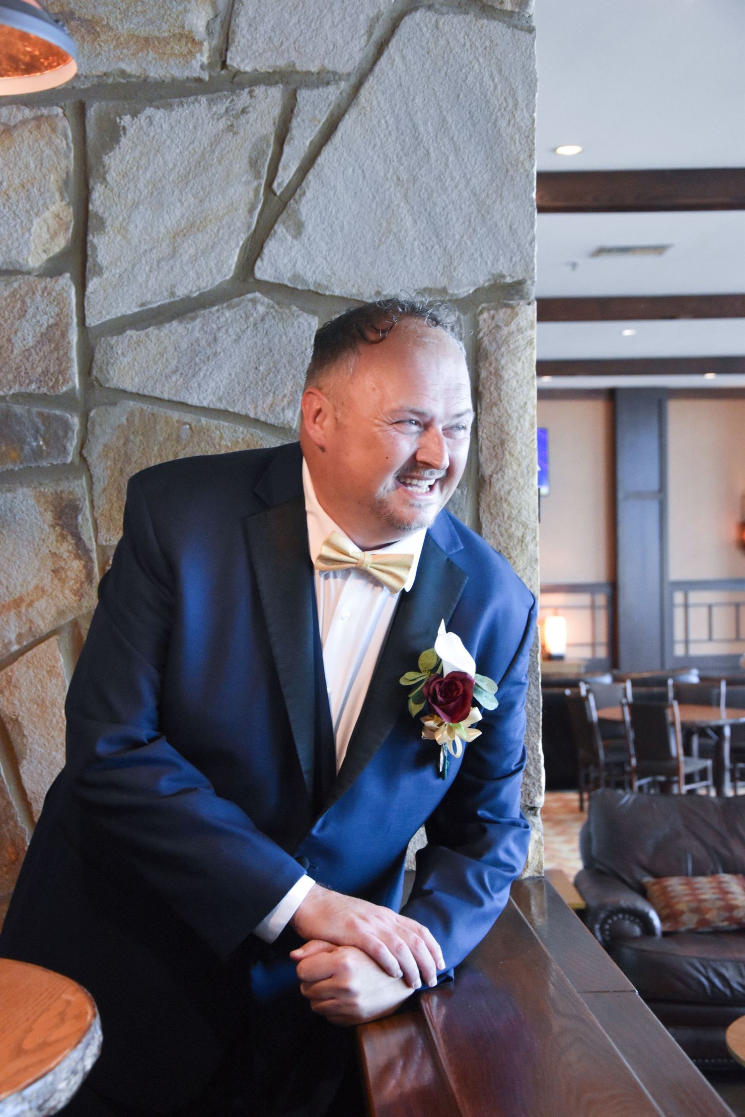 A person in a navy blue suit with a bow tie and boutonniere leans against a bar counter in an elegant venue.