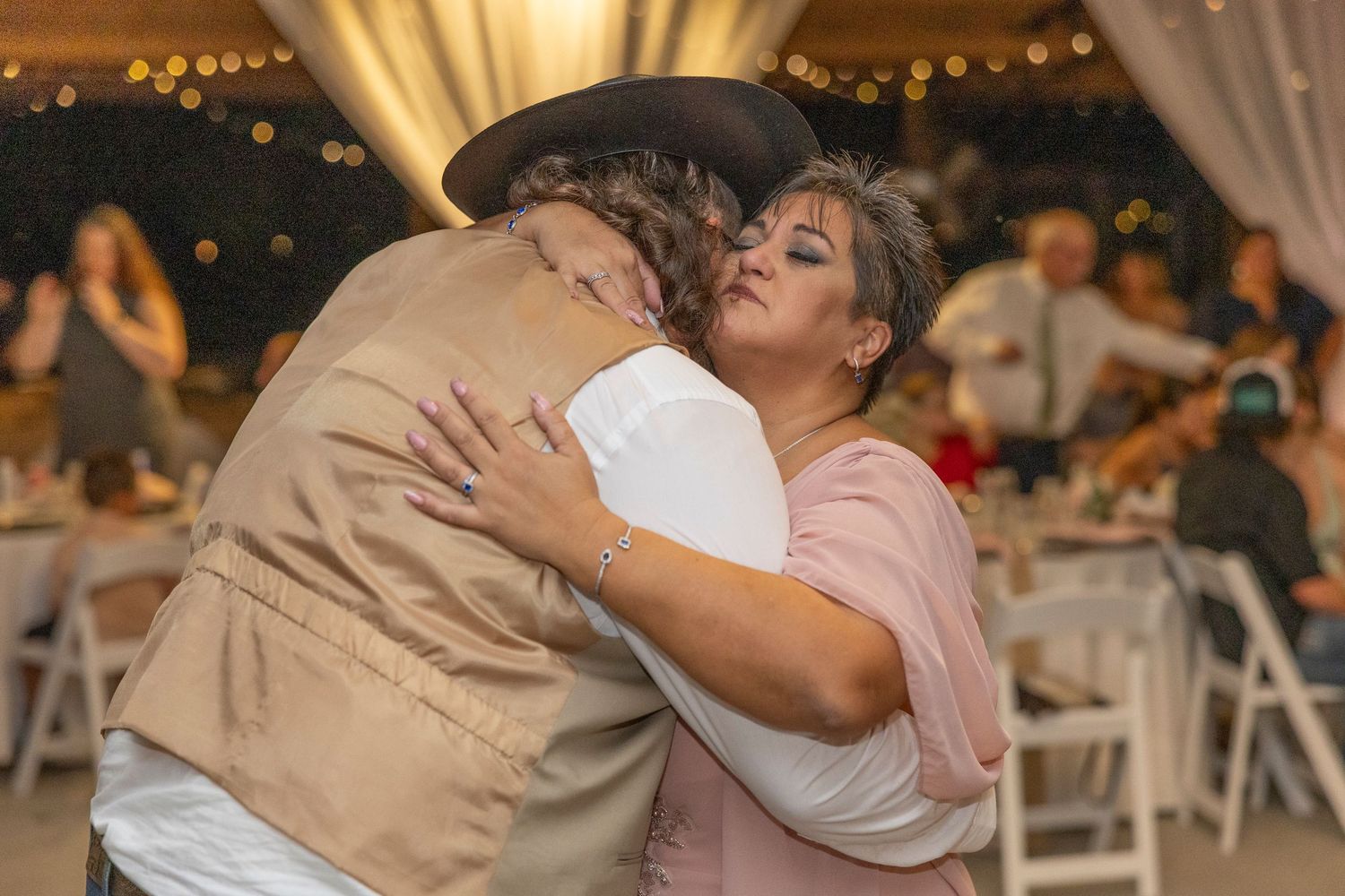 A couple shares an intimate dance embrace at a wedding reception with string lights and white decor in the background.