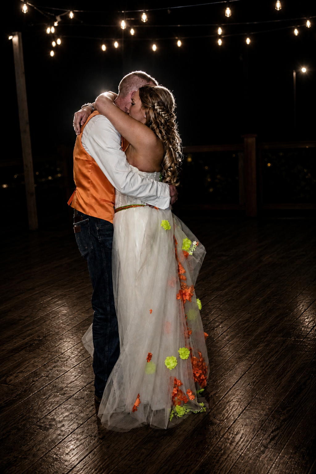 Bride and groom share first dance under string lights, her floral wedding dress glowing on a wooden dance floor.